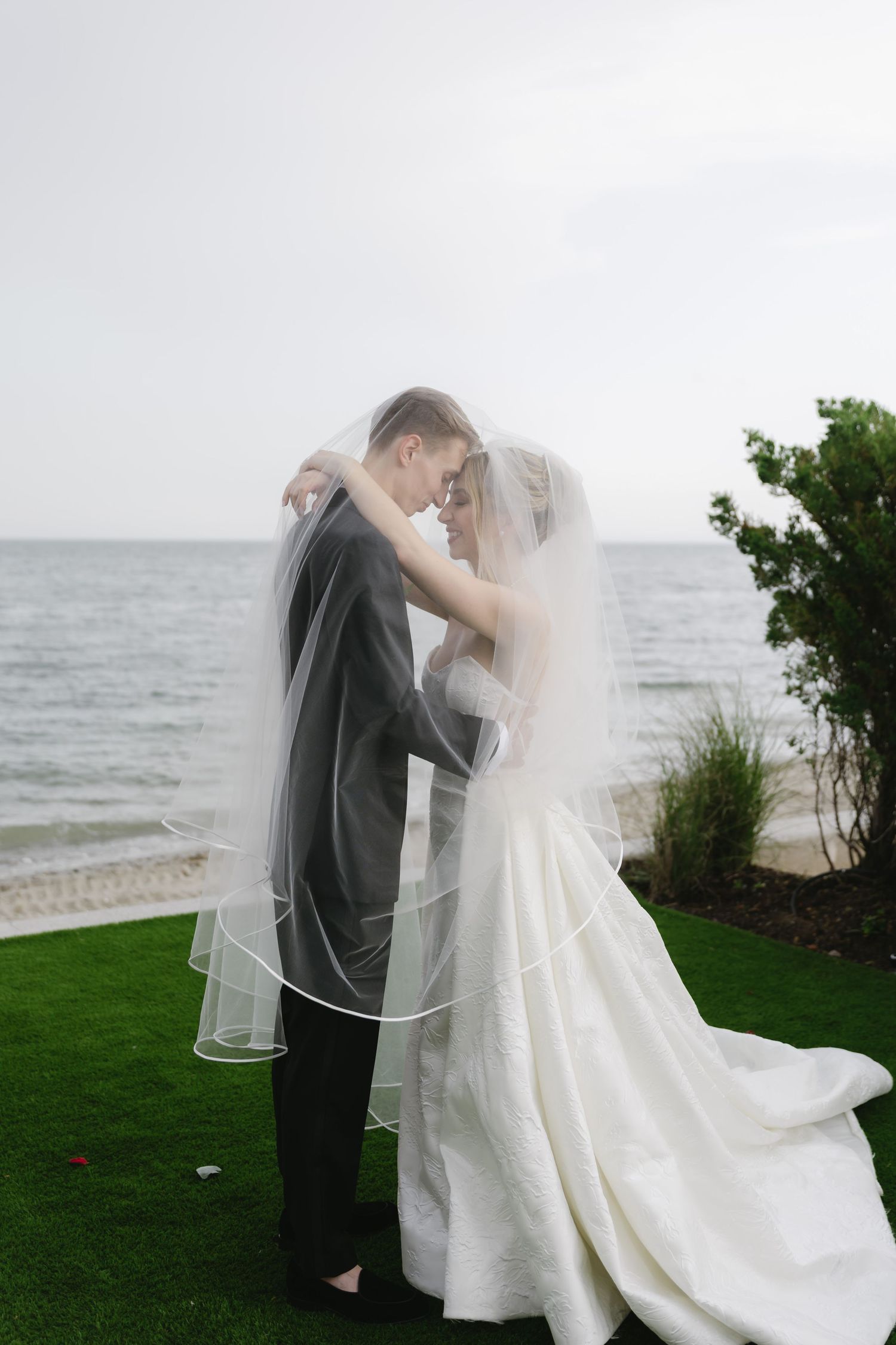 A romantic seaside wedding couple shares an intimate moment on a grassy lawn overlooking the ocean.