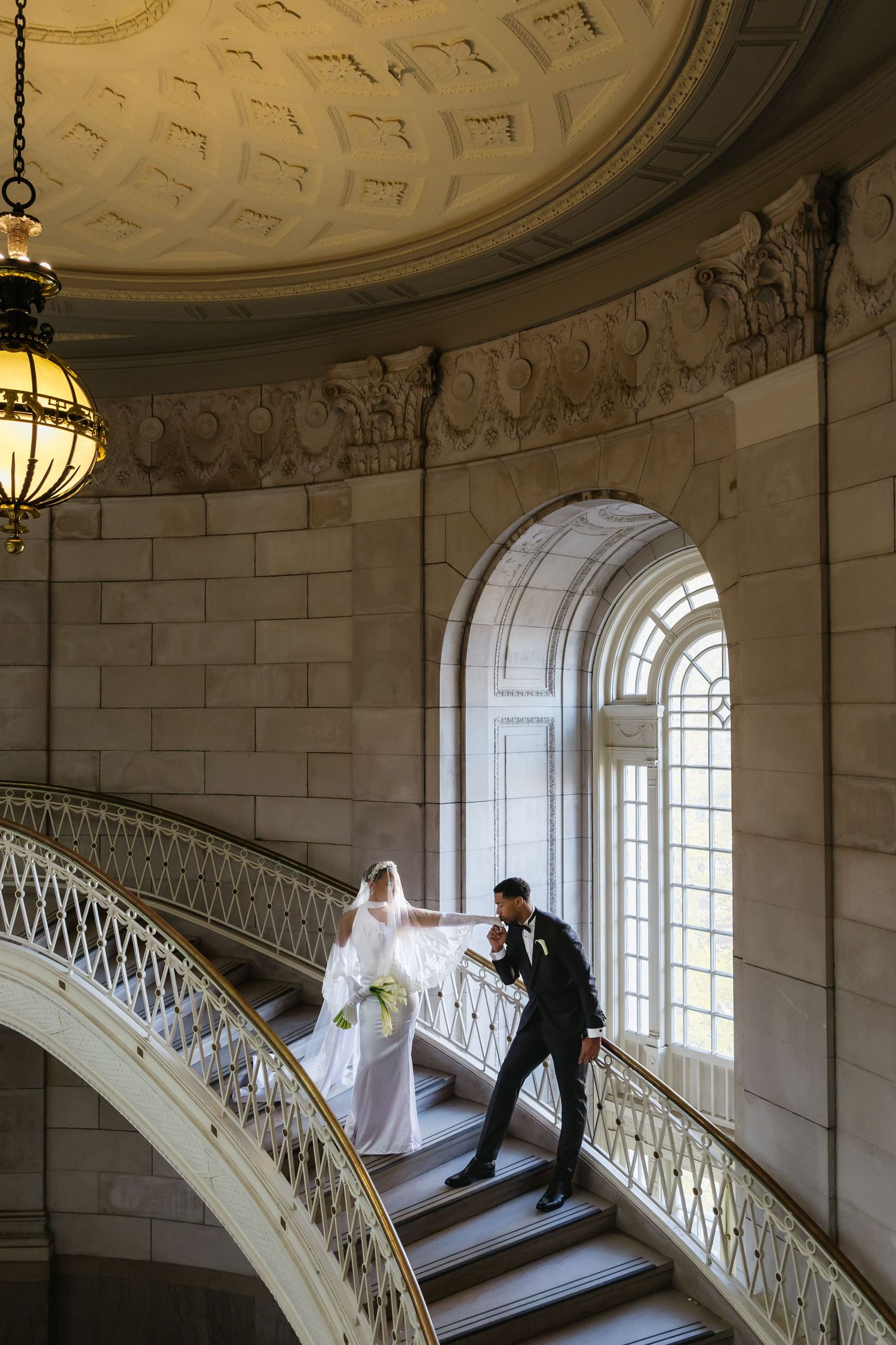 A couple in wedding attire poses on an elegant white marble spiral staircase with ornate railings and arched windows.