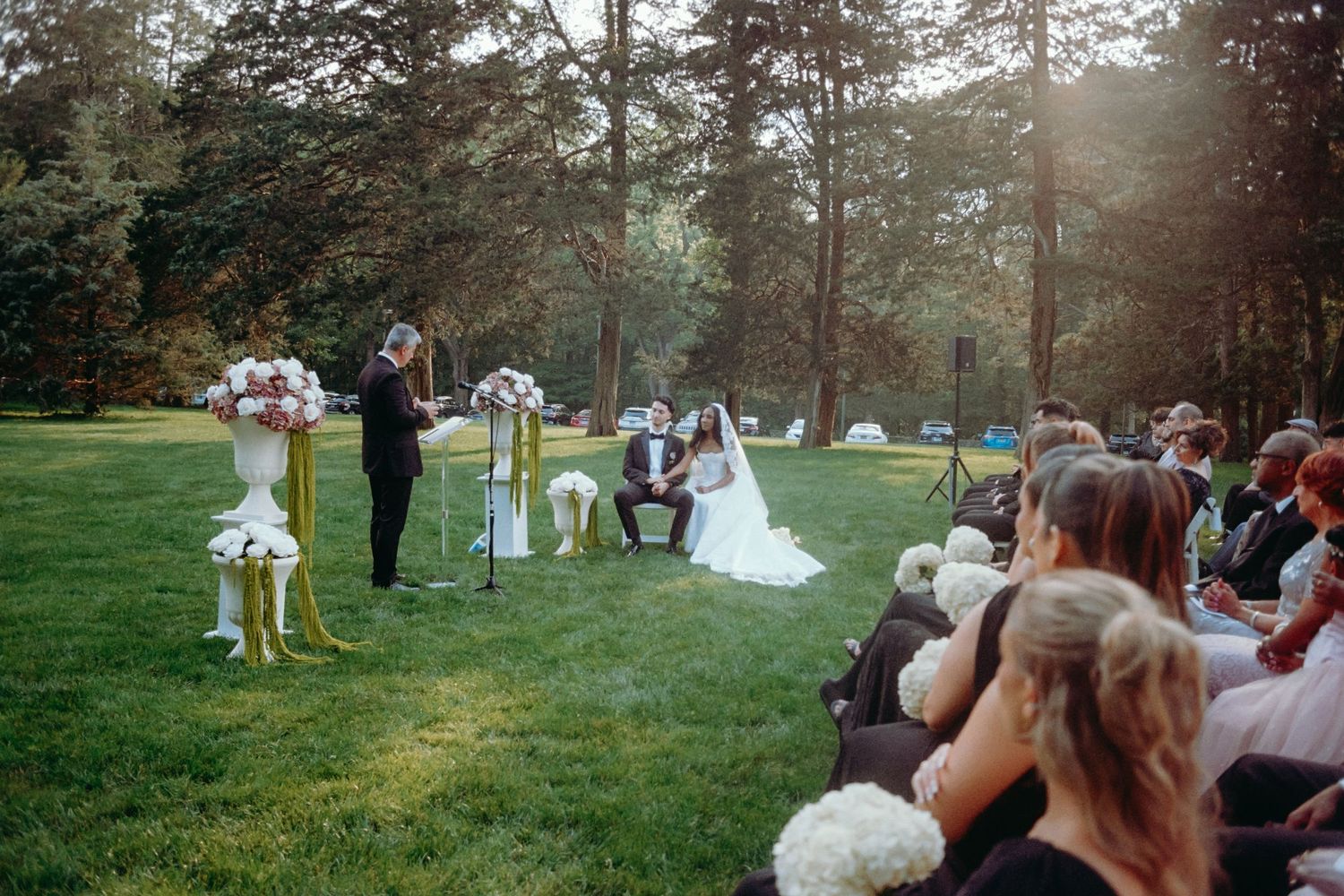 Outdoor wedding ceremony in a sunlit forest setting with guests seated on lawn.