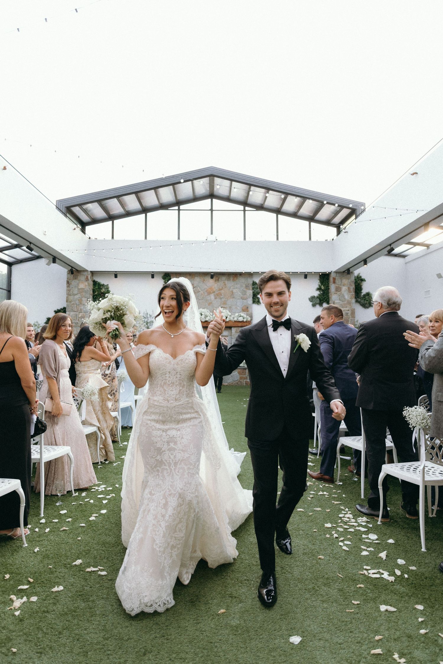Wedding party celebrates joyfully on a lawn with white flower petals scattered around a covered outdoor venue.