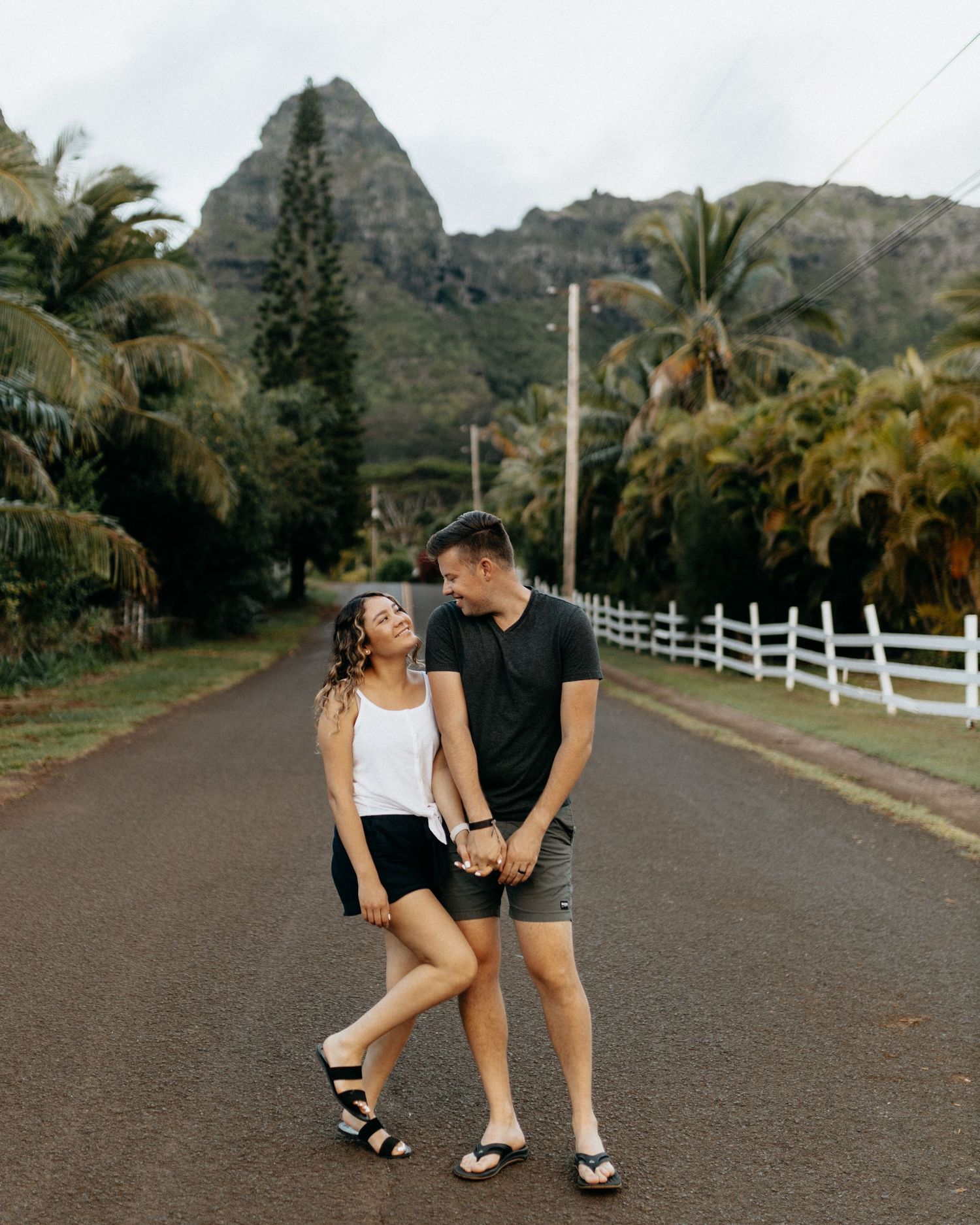 A couple walks together on a palm-lined road with dramatic mountain peaks rising in the background in Hawaii.