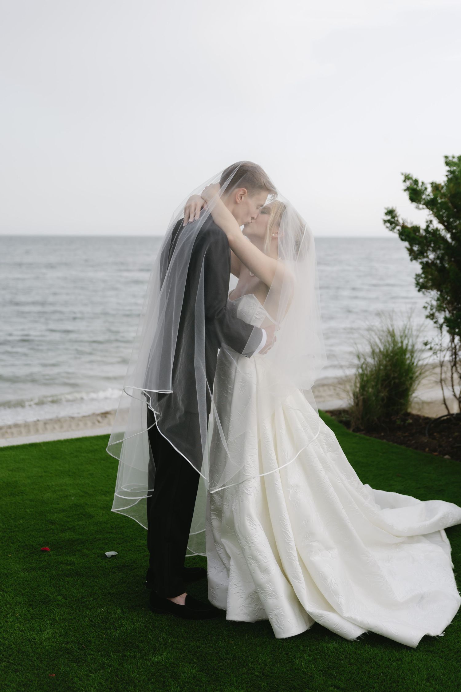 A romantic wedding couple embraces on a grassy cliff overlooking the ocean as the bride's veil flows in the breeze.