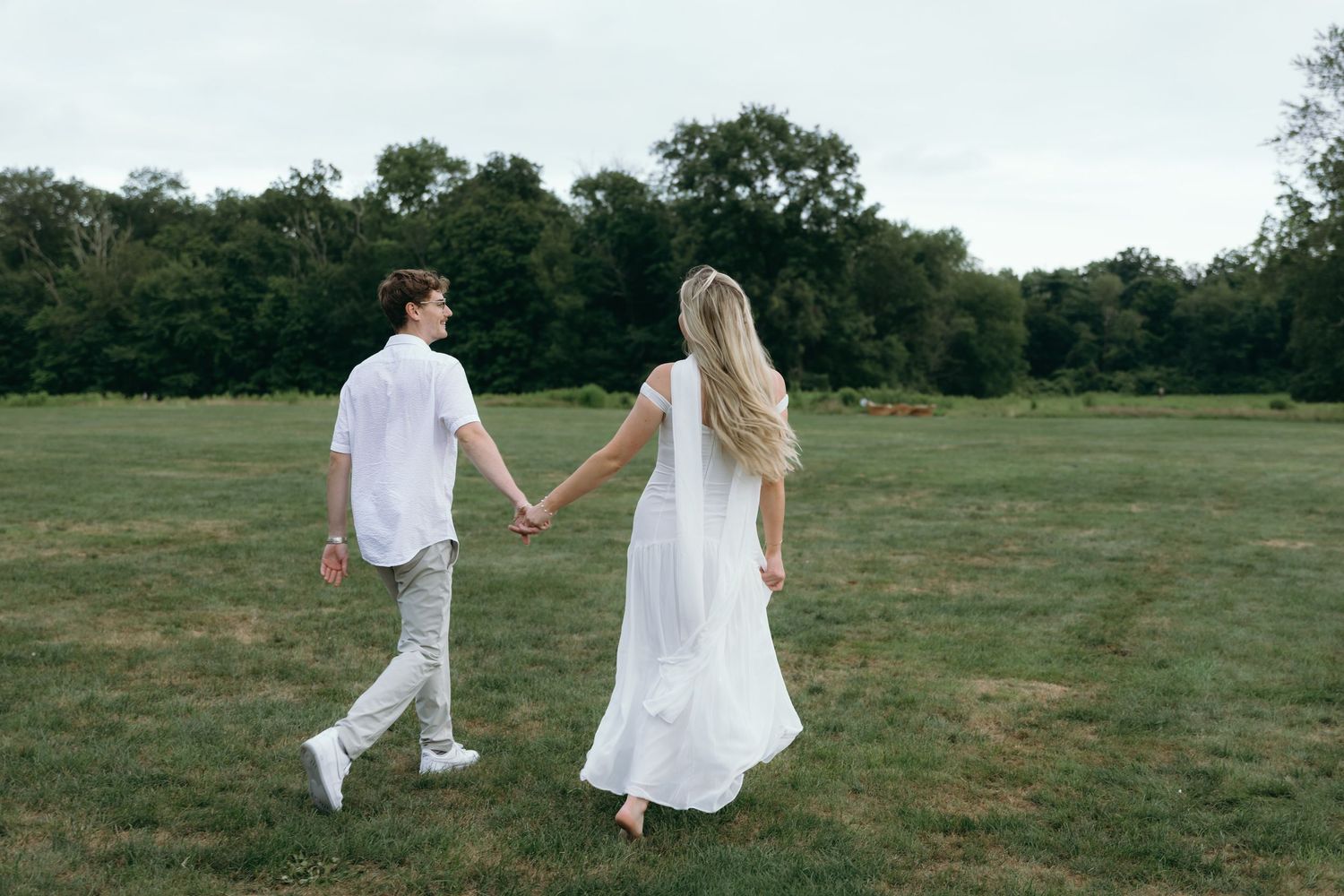 A couple in white outfits walks hand in hand through a green meadow on a sunny summer day.
