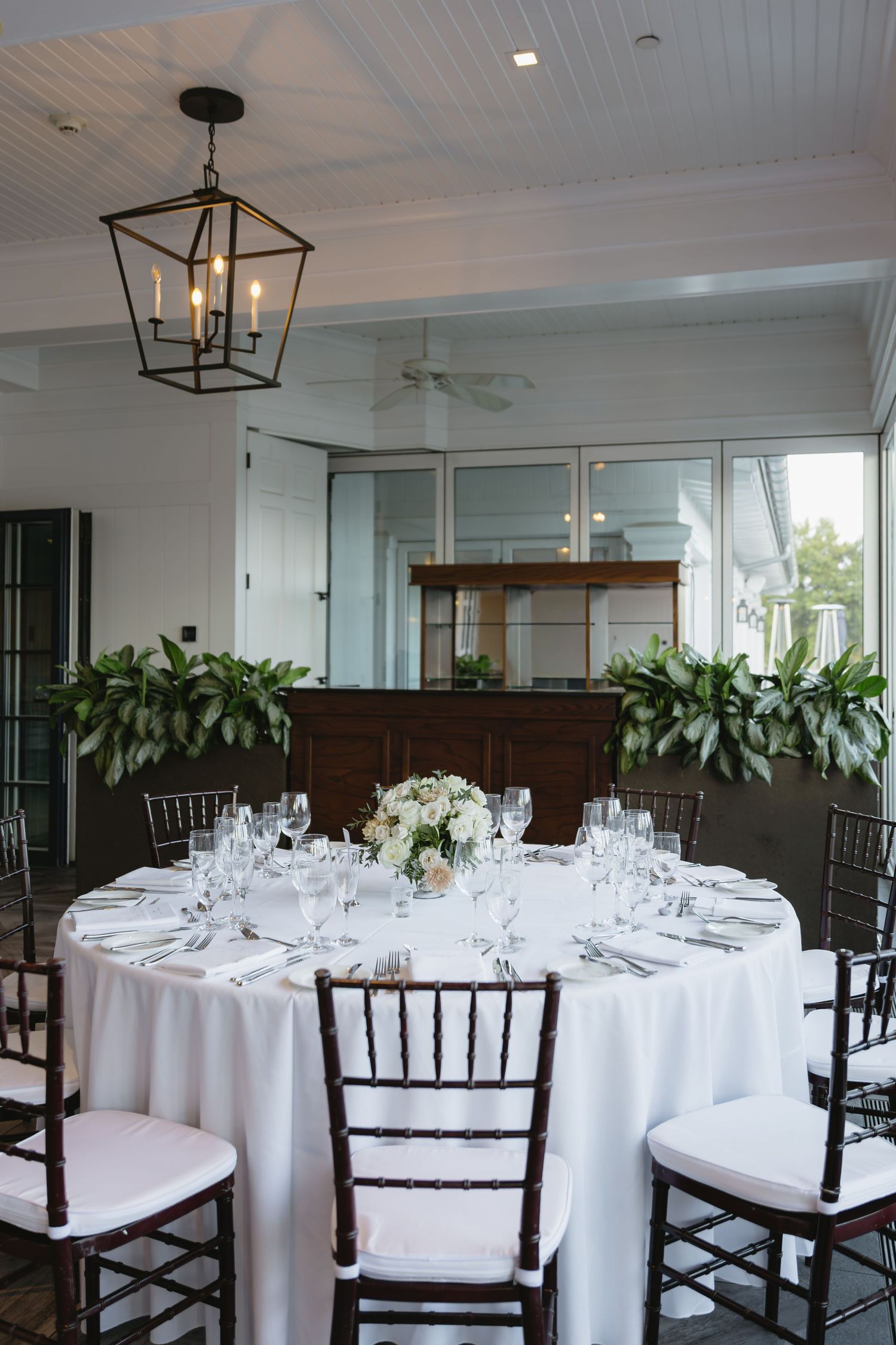 Elegant wedding reception venue with round tables, white linens, dark wood chairs, and modern lantern lighting fixtures.