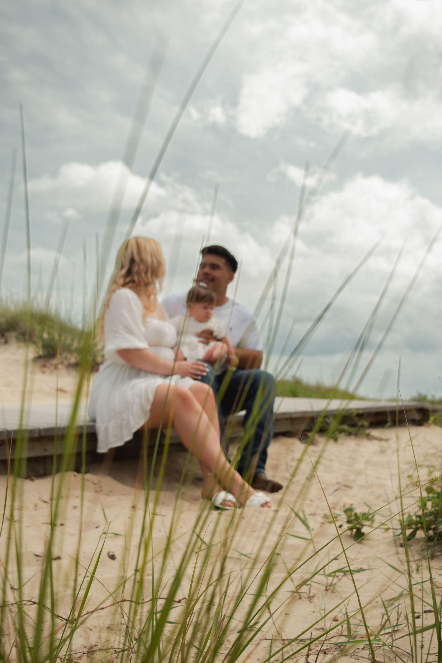 A family of three sits together on sandy beach dunes surrounded by tall grass under a cloudy sky.