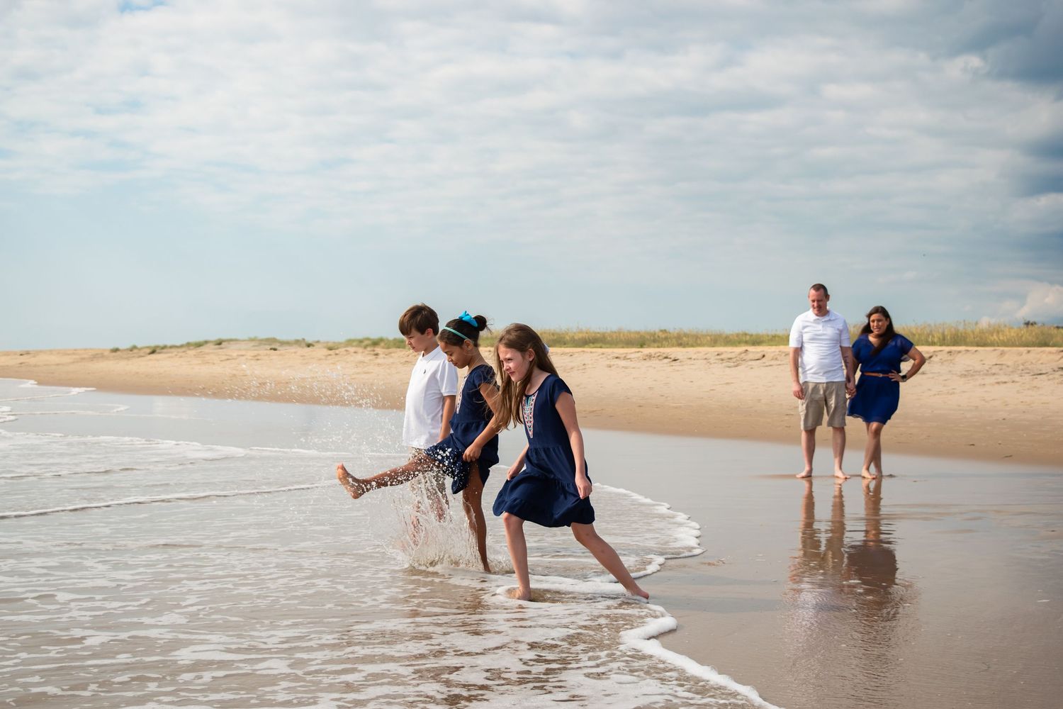 Family enjoying a beach day, with two children splashing in ocean waves while parents watch from the sandy shore.
