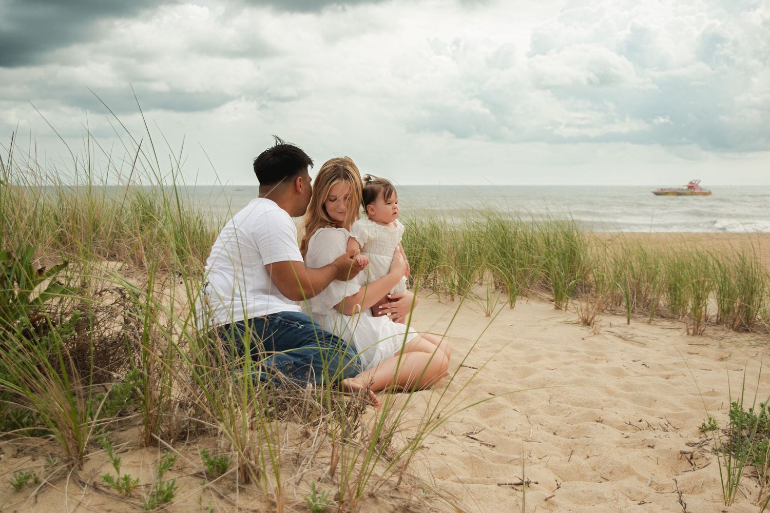 Family sitting together on sandy beach dunes with ocean and cloudy sky in background.