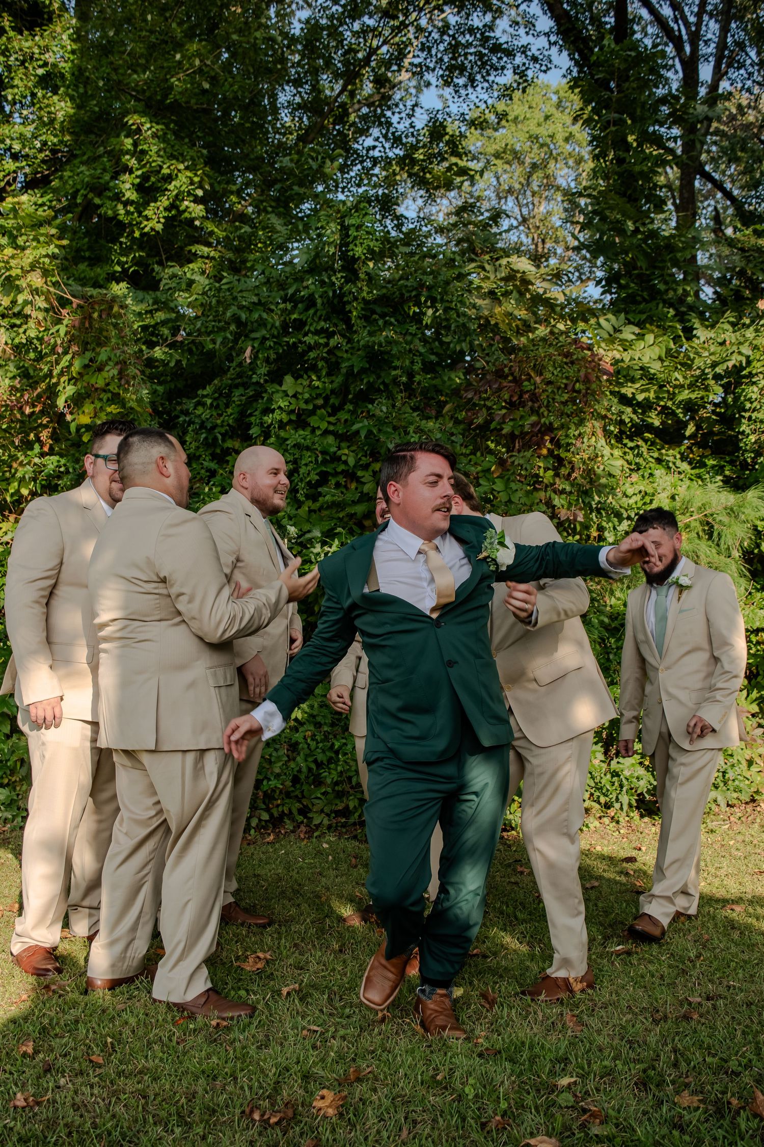 Wedding party in tan suits and green attire laugh together while posing outdoors in a garden setting.