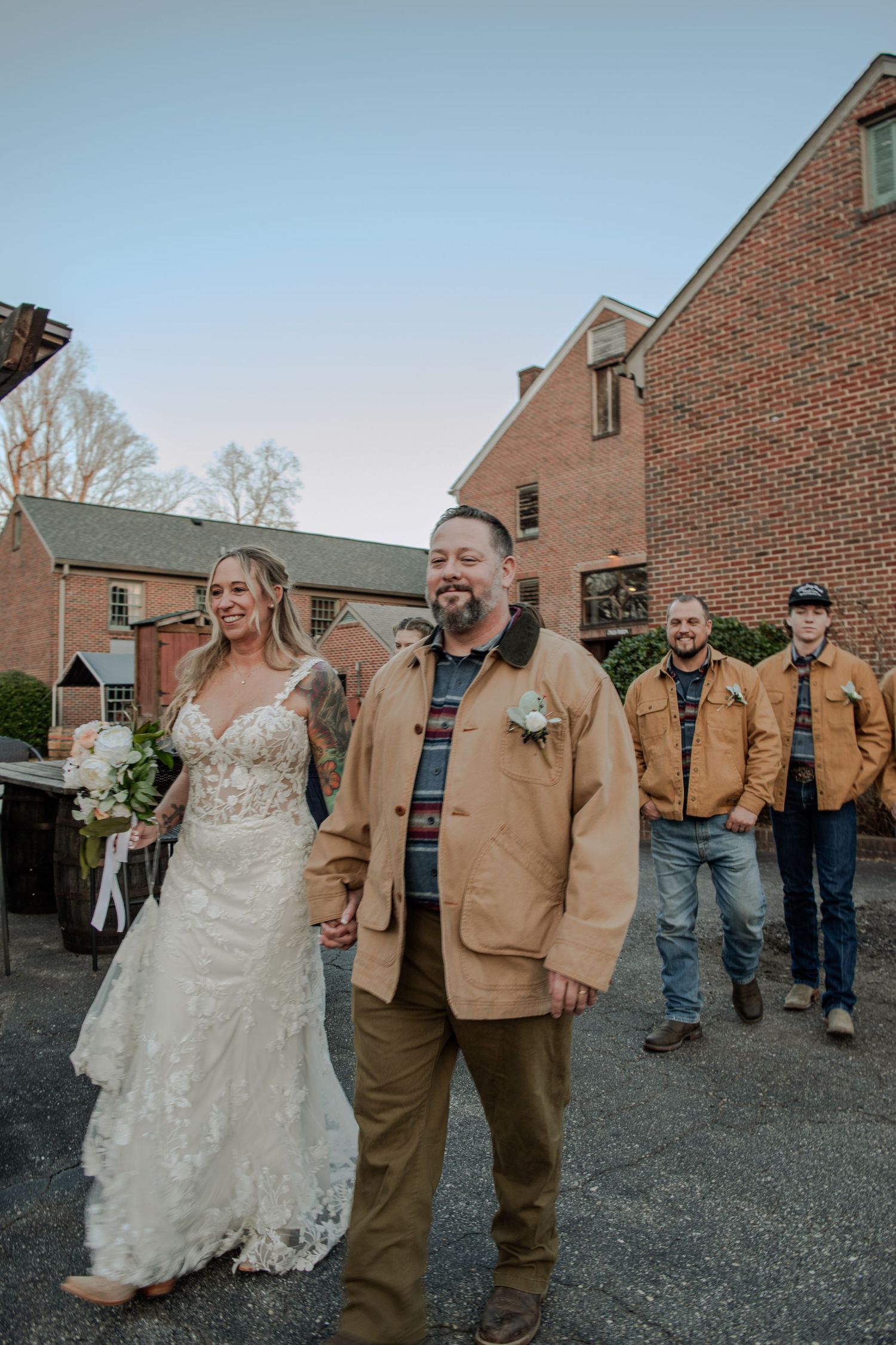 A wedding party walks together on a gravel path near brick buildings in matching tan jackets.