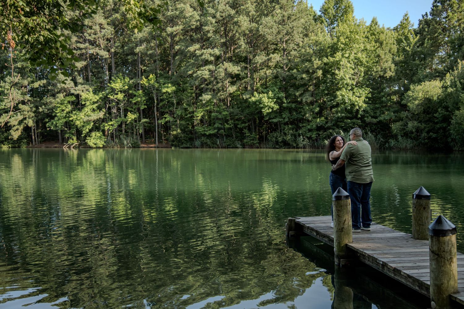 Couple embracing on a wooden dock overlooking a calm, reflective green lake surrounded by lush forest trees.