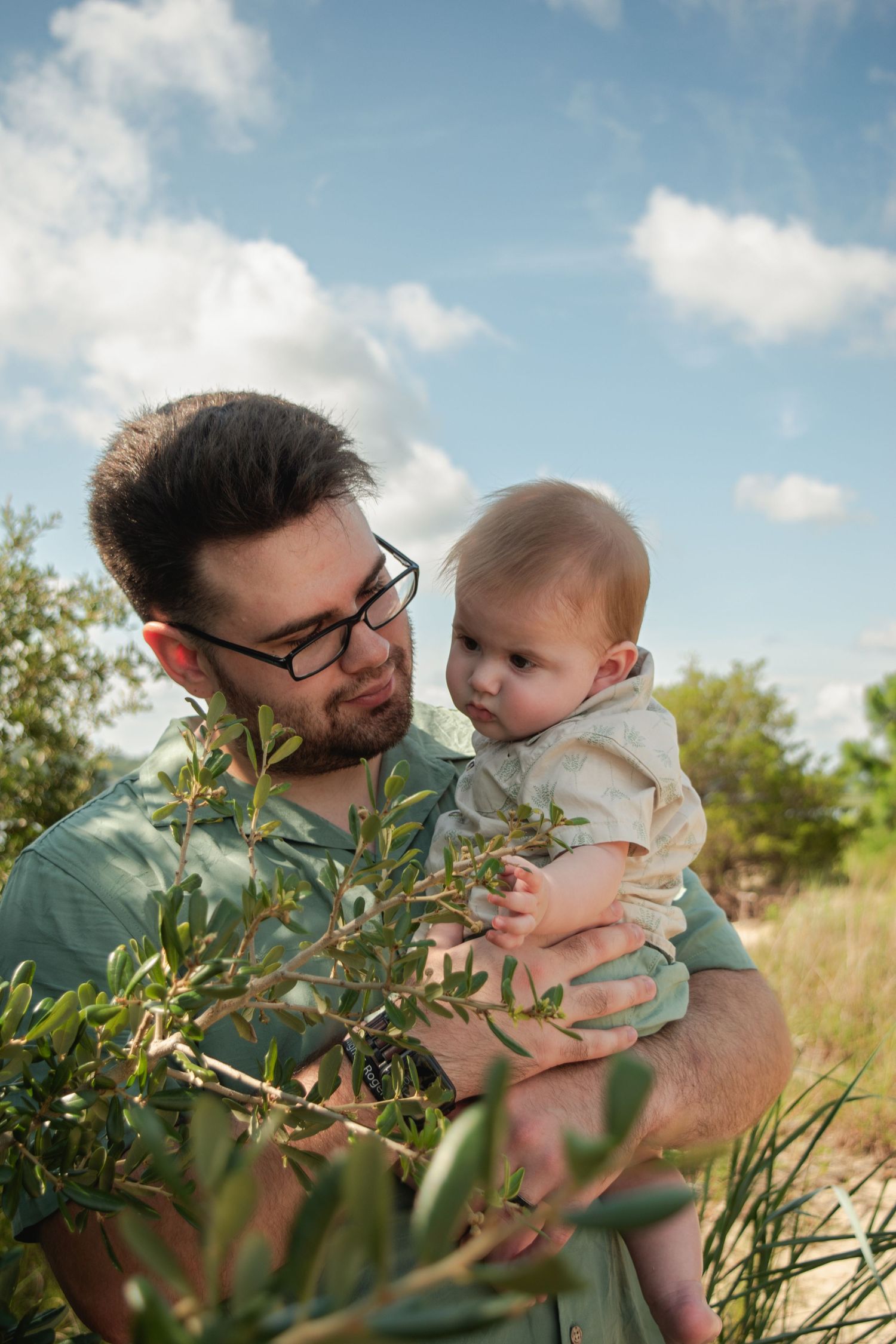 Father with glasses holds baby outdoors near green shrubs under a partly cloudy sky.
