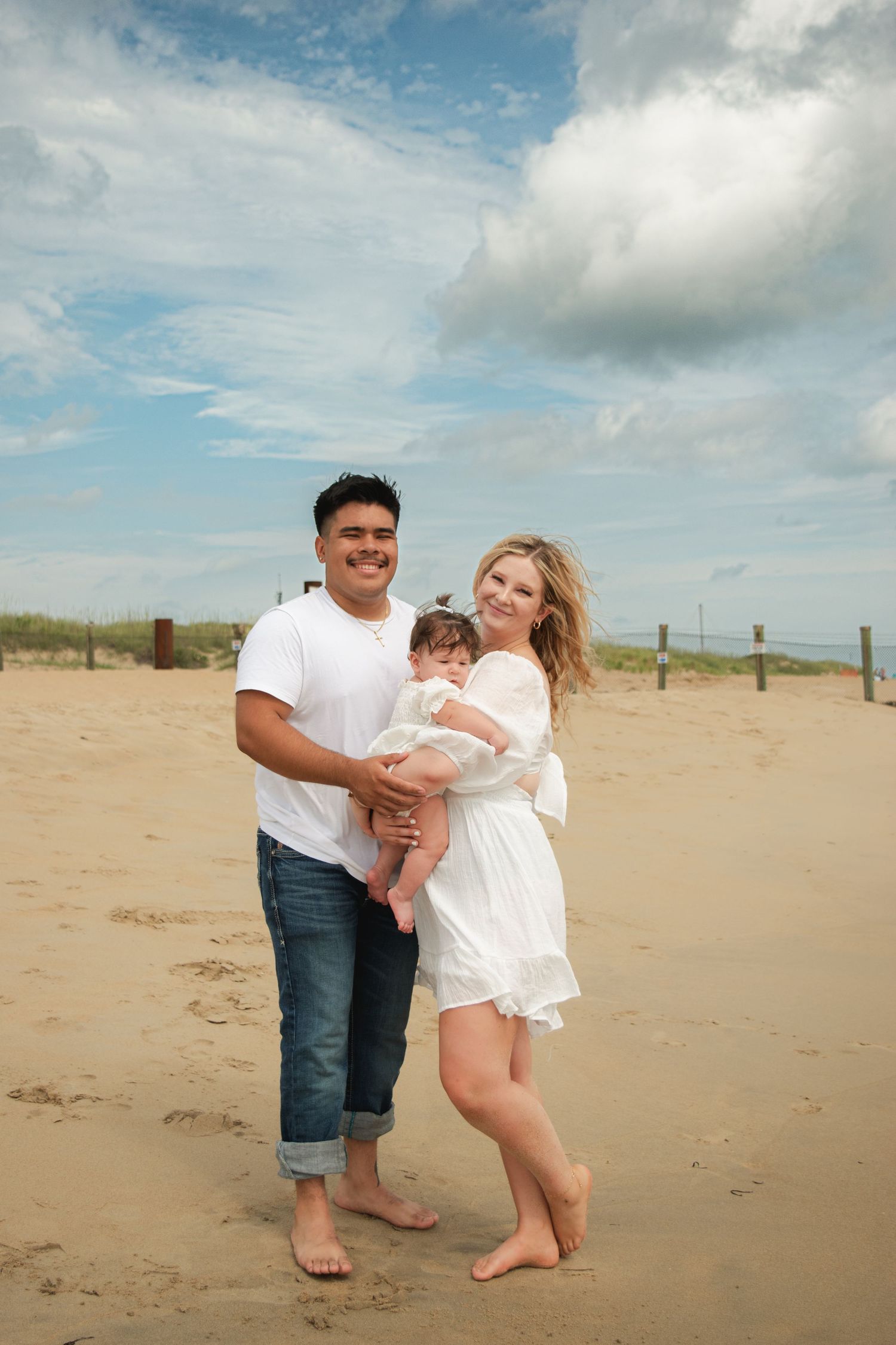 A smiling couple stands barefoot on the beach holding their baby girl, posing together under a partly cloudy sky.