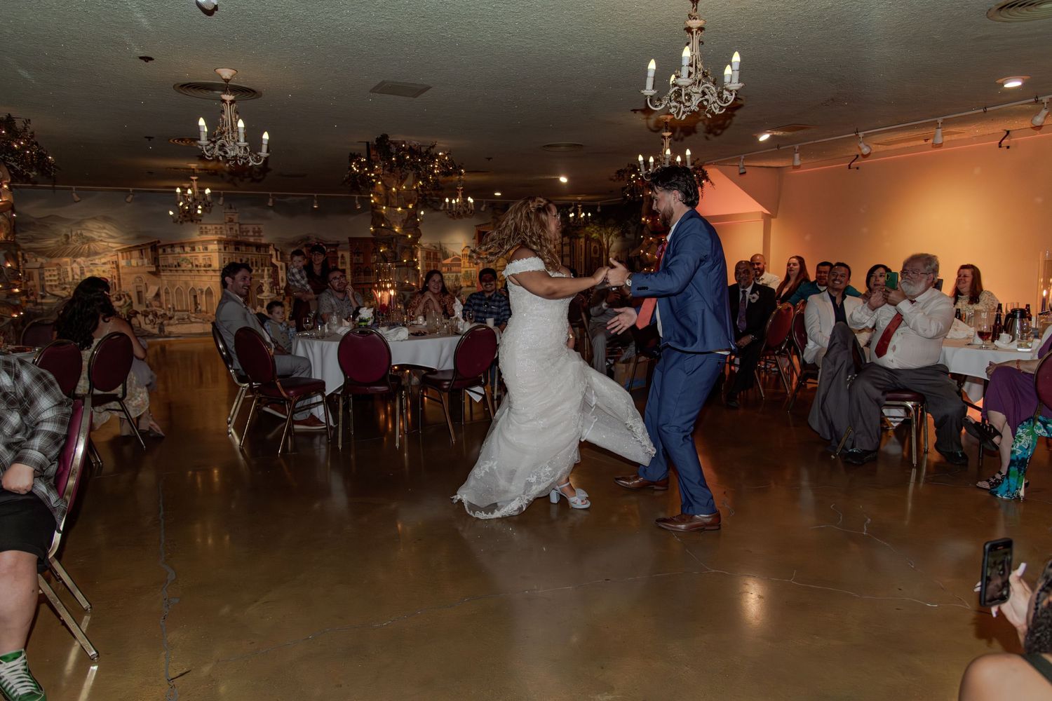 Bride and groom share their first dance at a romantic wedding reception in an elegant stone-walled venue with chandelier lighting.