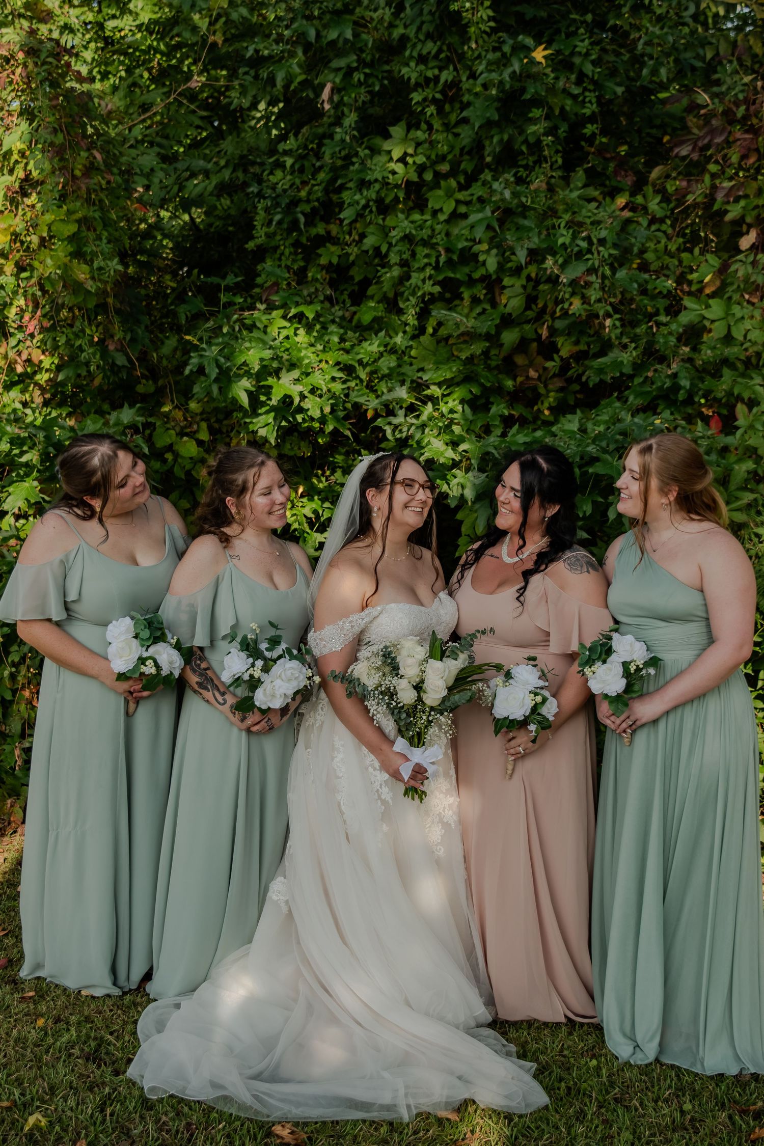 Bridesmaids in sage green and blush dresses pose with bride in white gown during outdoor wedding photos.