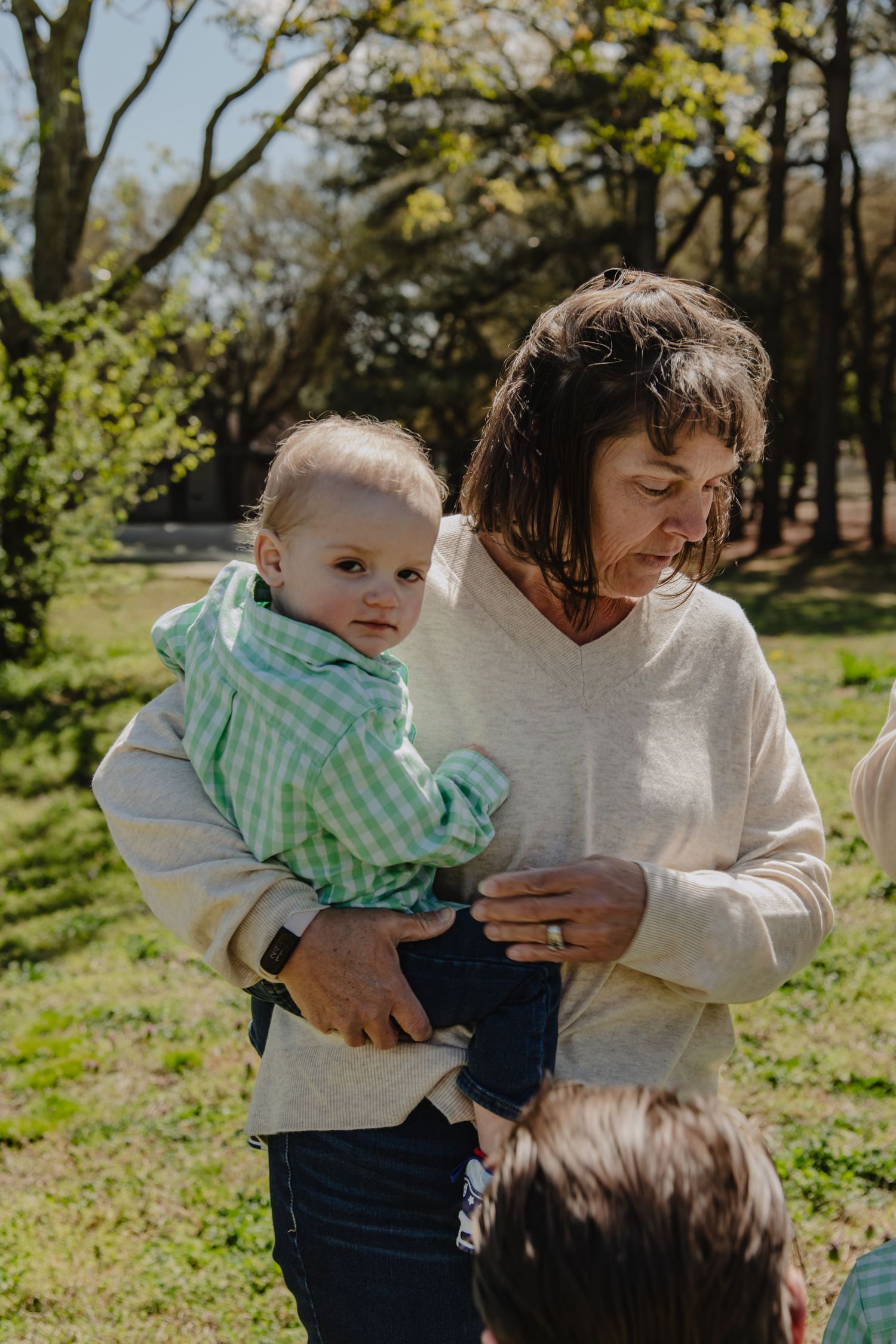 A woman in a cream sweater holds a baby in a green gingham shirt outdoors in a sunny park setting.