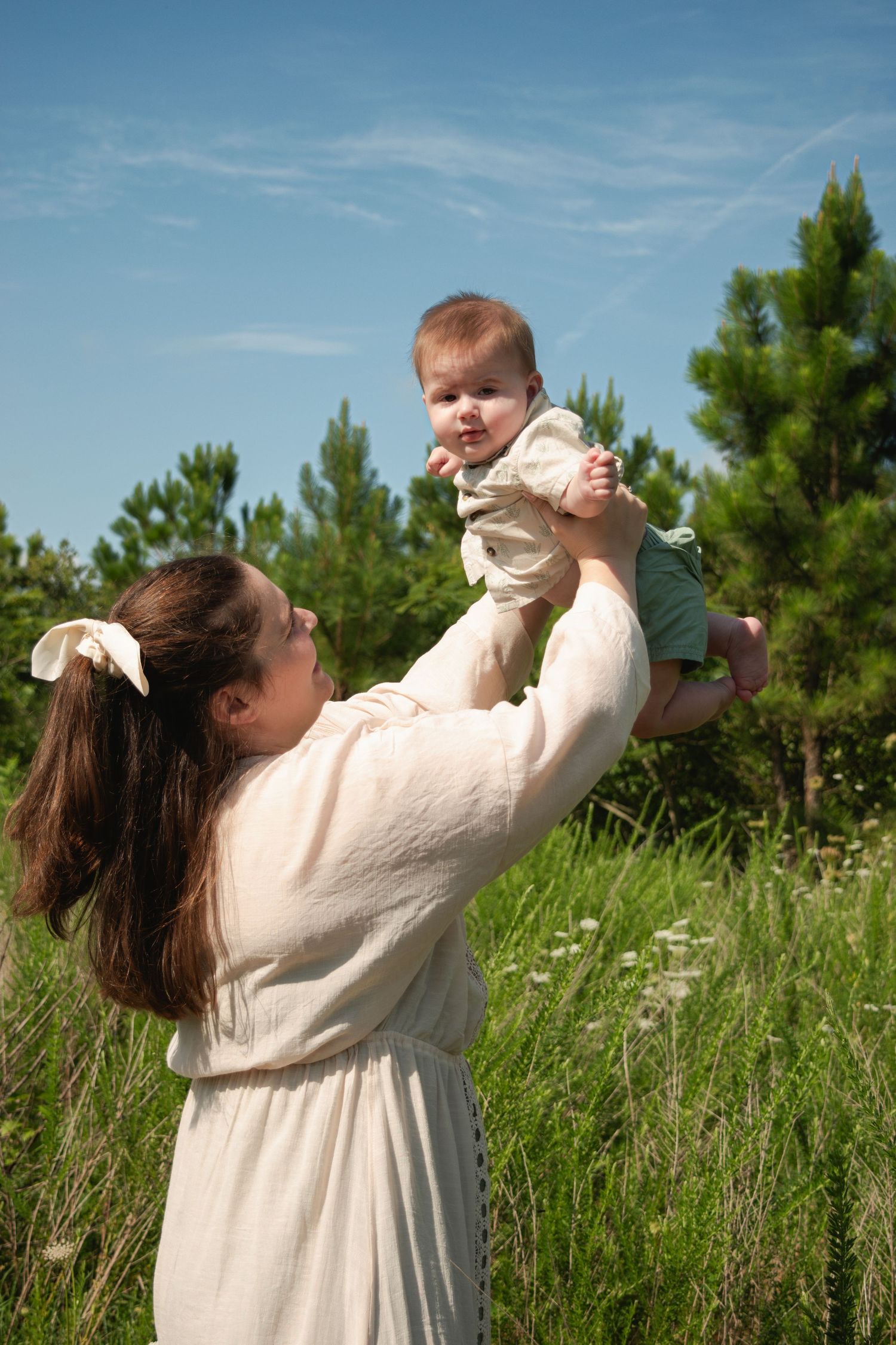 A mother lifts her smiling baby overhead in a sunny green field with trees and blue sky in the background.