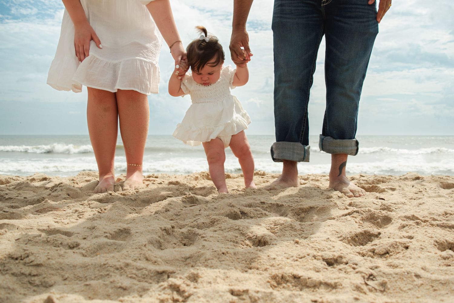 Parents holding baby's hands while learning to walk on sandy beach with ocean waves in background.