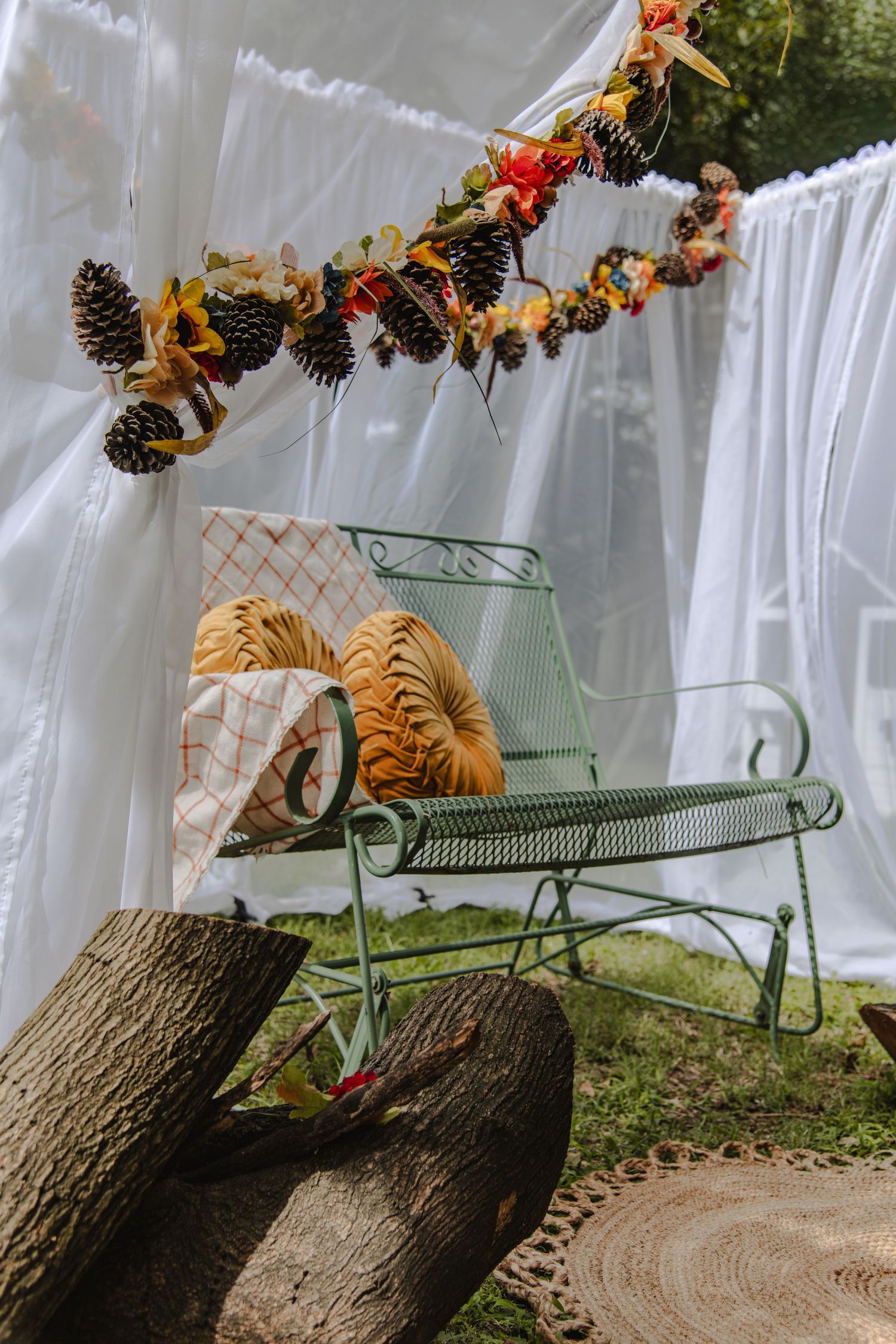 Pine cones and autumn leaves decorate a rustic holiday display with white curtains and green sleigh in outdoor setting.
