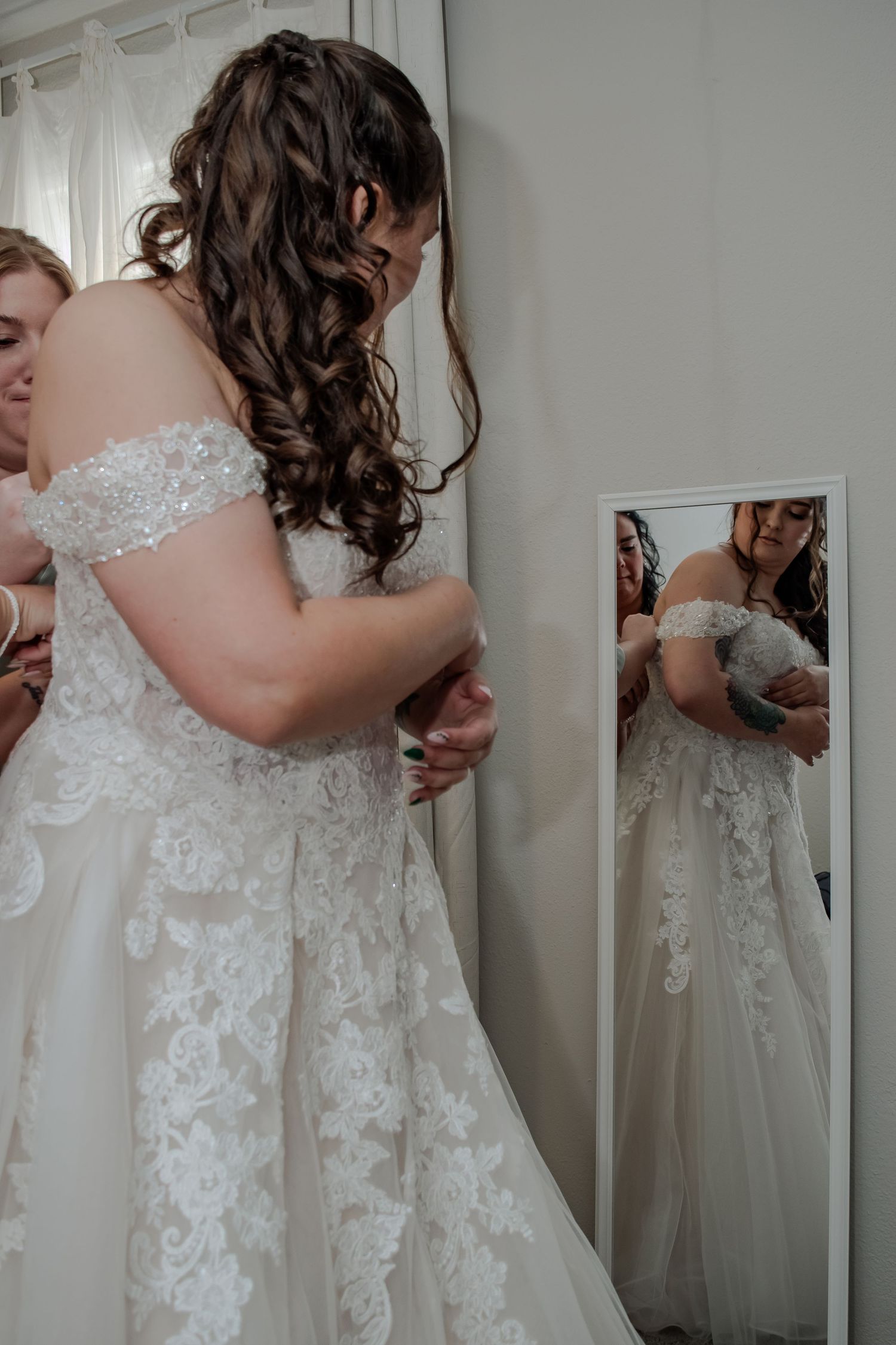 A bride in an elegant lace wedding gown with flowing curls gets ready while looking in a full-length mirror.