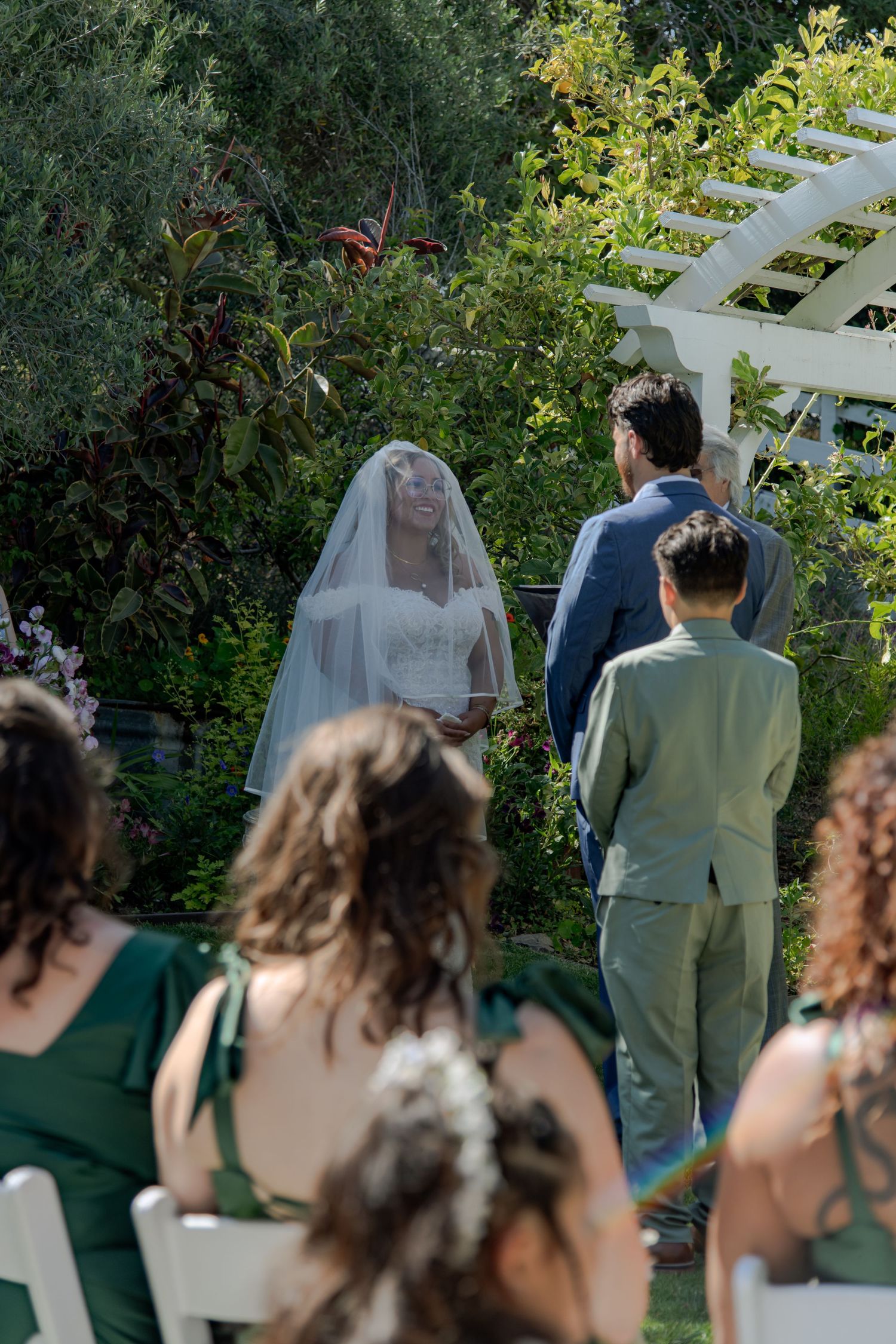 Wedding ceremony participants stand in a garden setting with white lattice and green foliage in the background.