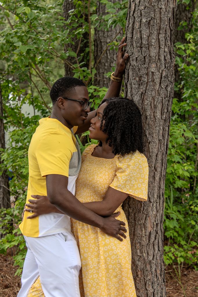 A couple in matching yellow outfits shares a romantic moment leaning against a tree in a lush green forest.