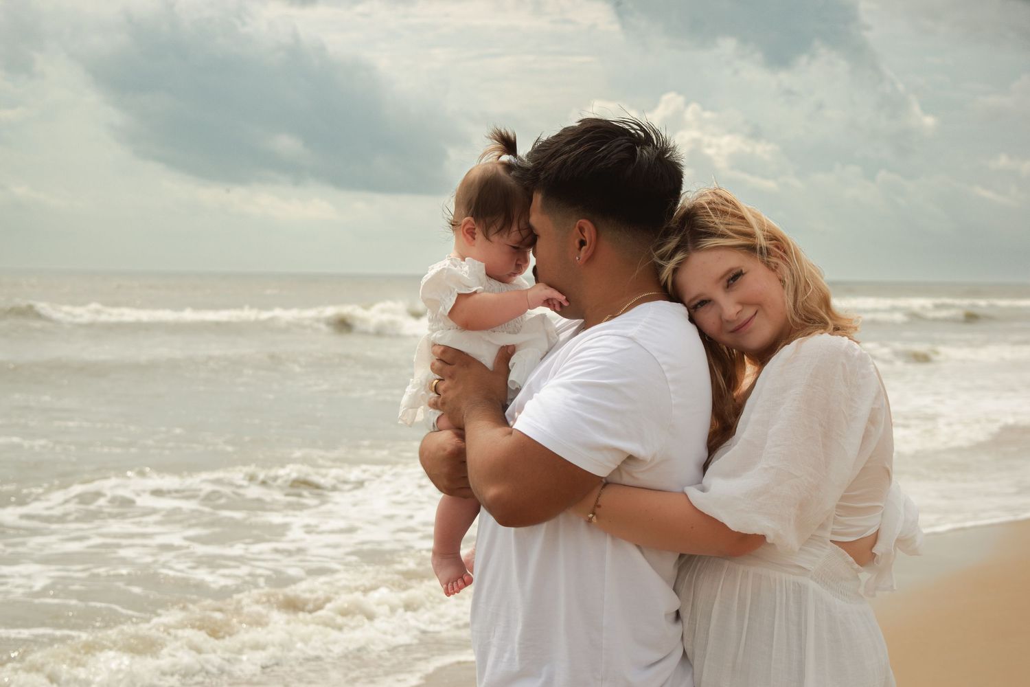 Young family with baby enjoying a beach day, man holding infant while woman leans on his shoulder.
