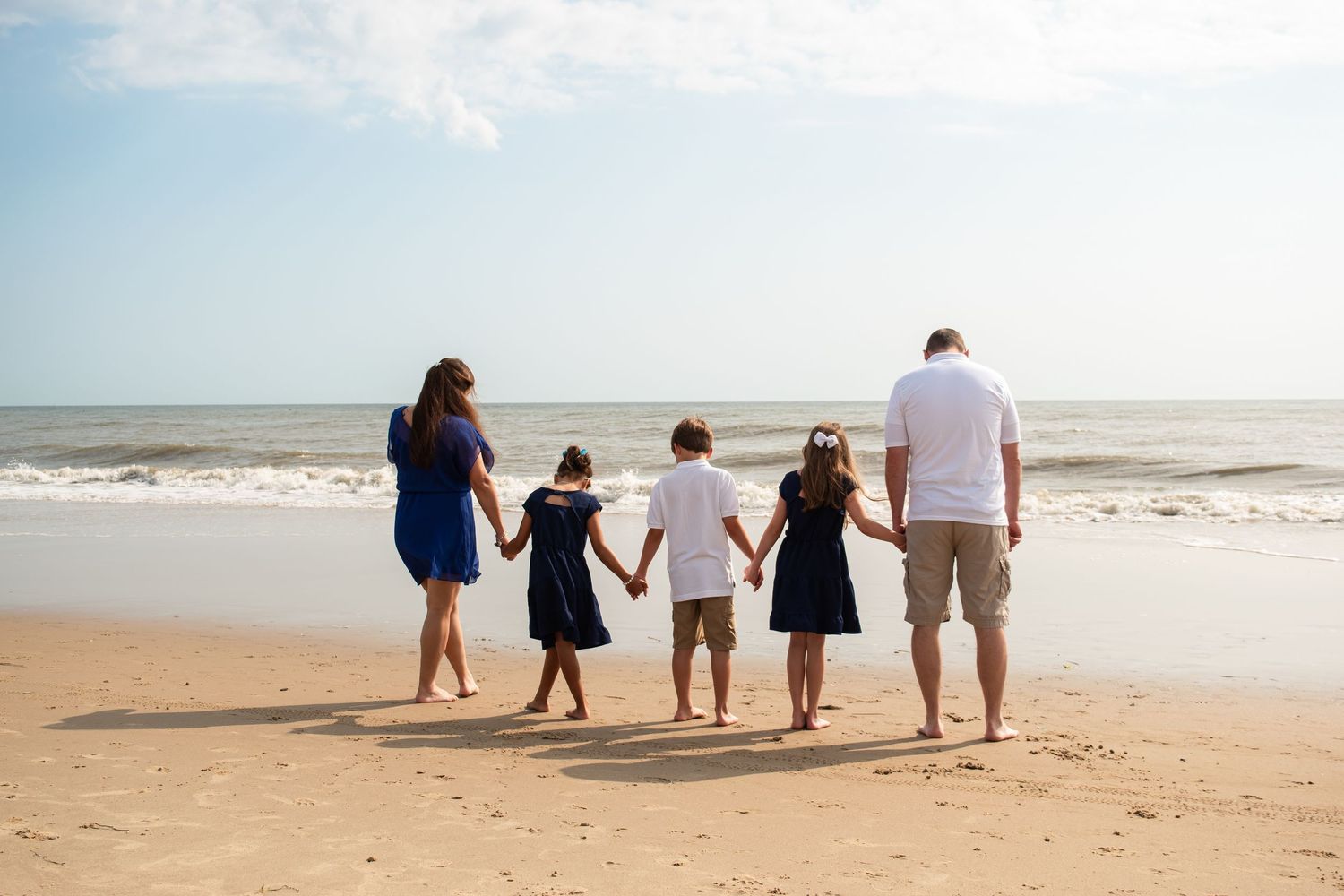 Family of five holding hands on a sandy beach, gazing at ocean waves under a clear blue sky.