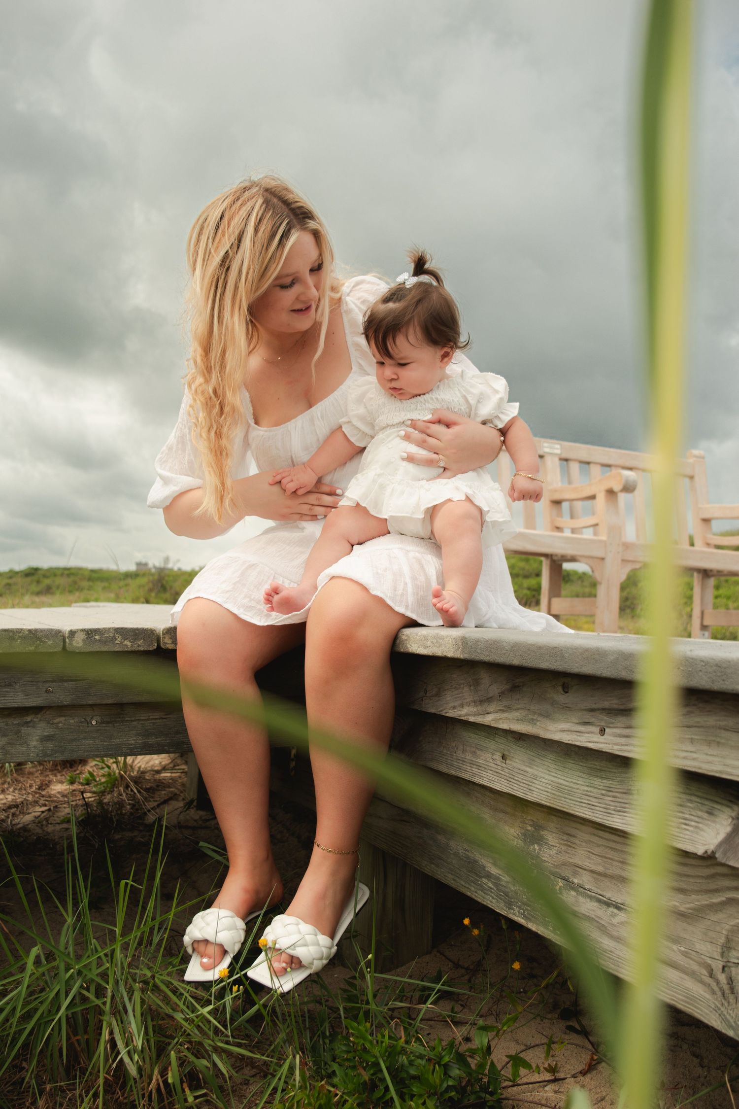 Blonde mother in white dress sitting on wooden bridge holding infant daughter outdoors.