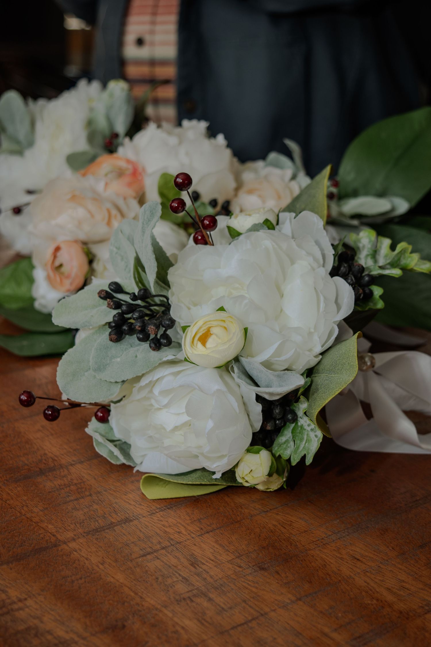 A stunning bridal bouquet with white roses, peonies, and dark berries rests on a wooden surface.