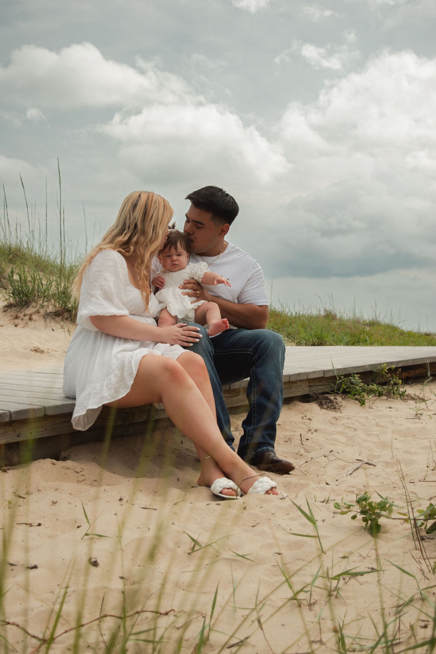 A young family sits together on a sandy beach boardwalk, parents lovingly attending to their baby girl under a cloudy sky.