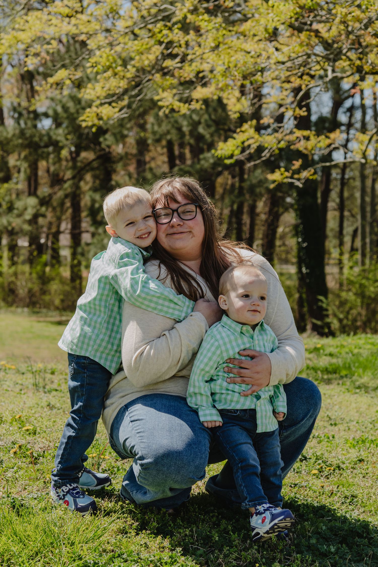 A mother crouches outdoors hugging two young boys in matching green plaid shirts, surrounded by lush green trees.