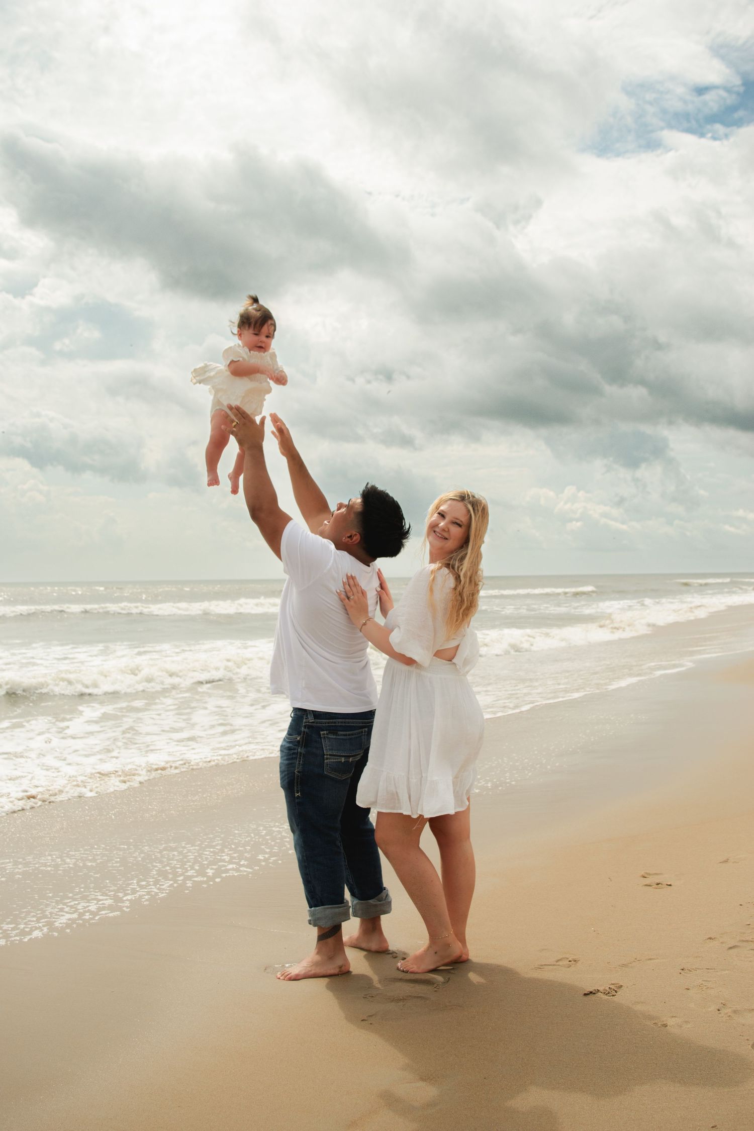 Happy family on beach, father lifting baby in air while mother watches, ocean waves in background.