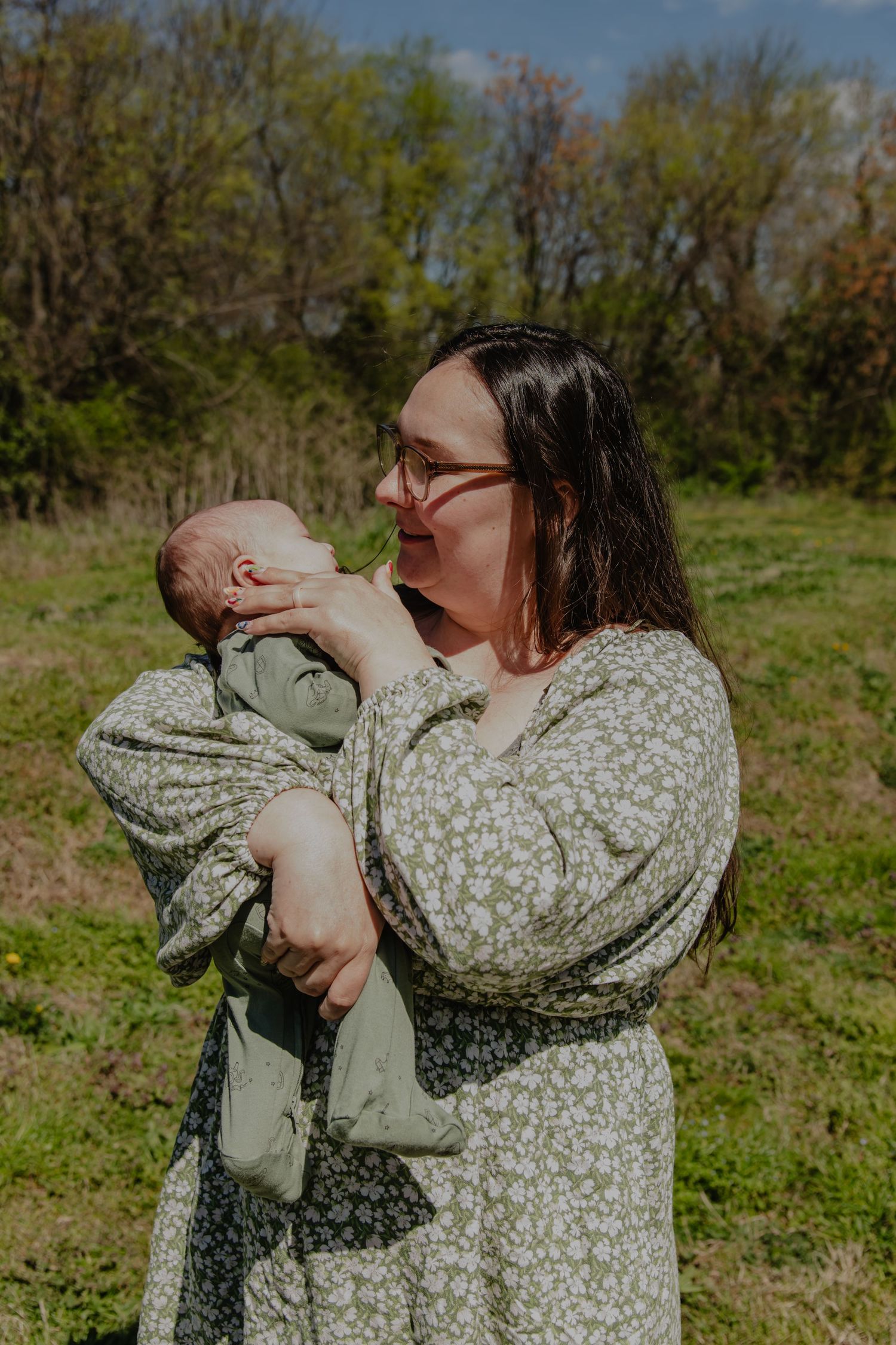 A mother in a floral green dress holds her newborn baby outdoors in a sunny green field.