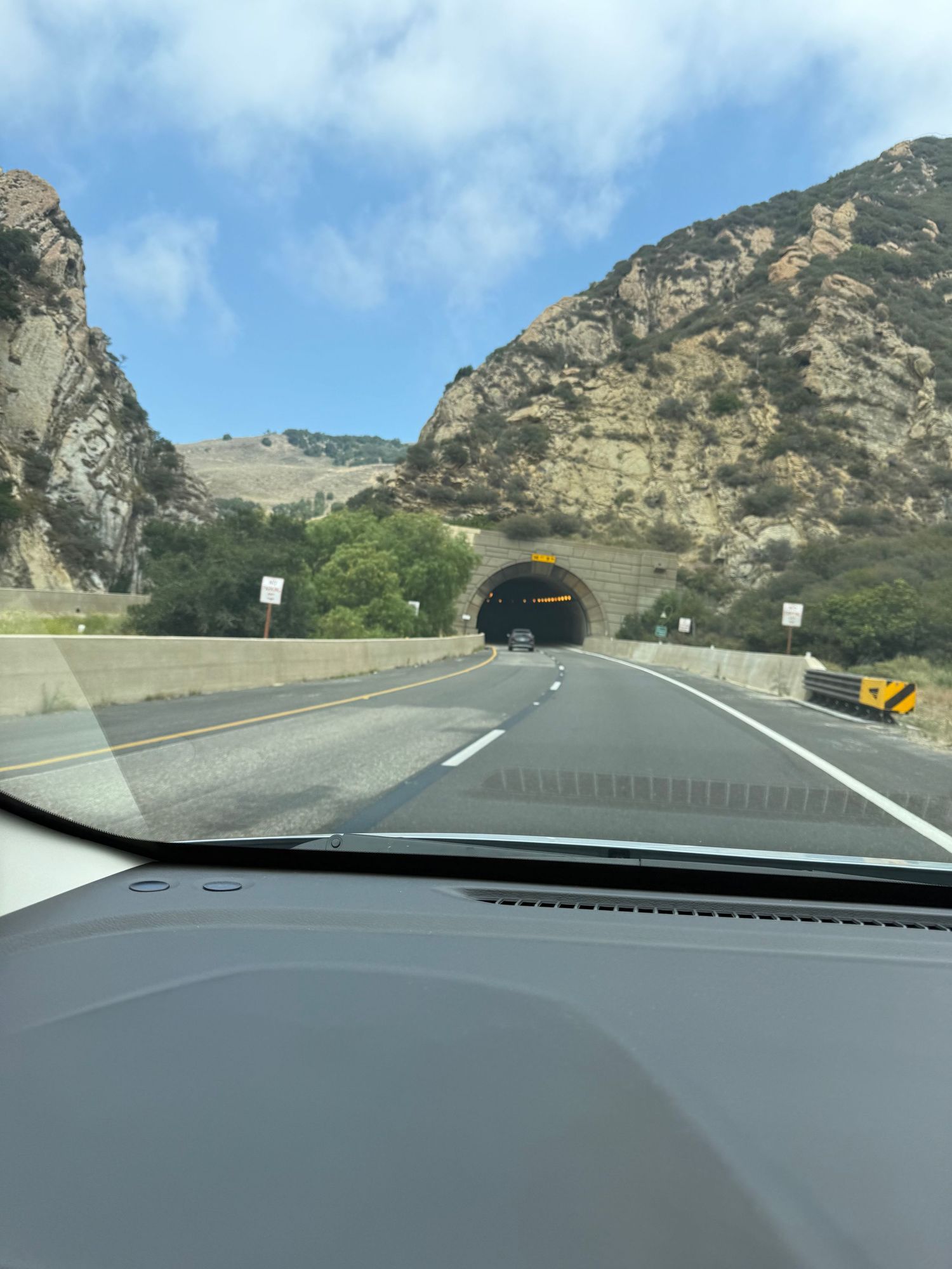 Car approaches tunnel entrance carved through rocky mountain pass on sunny day with blue skies visible above.