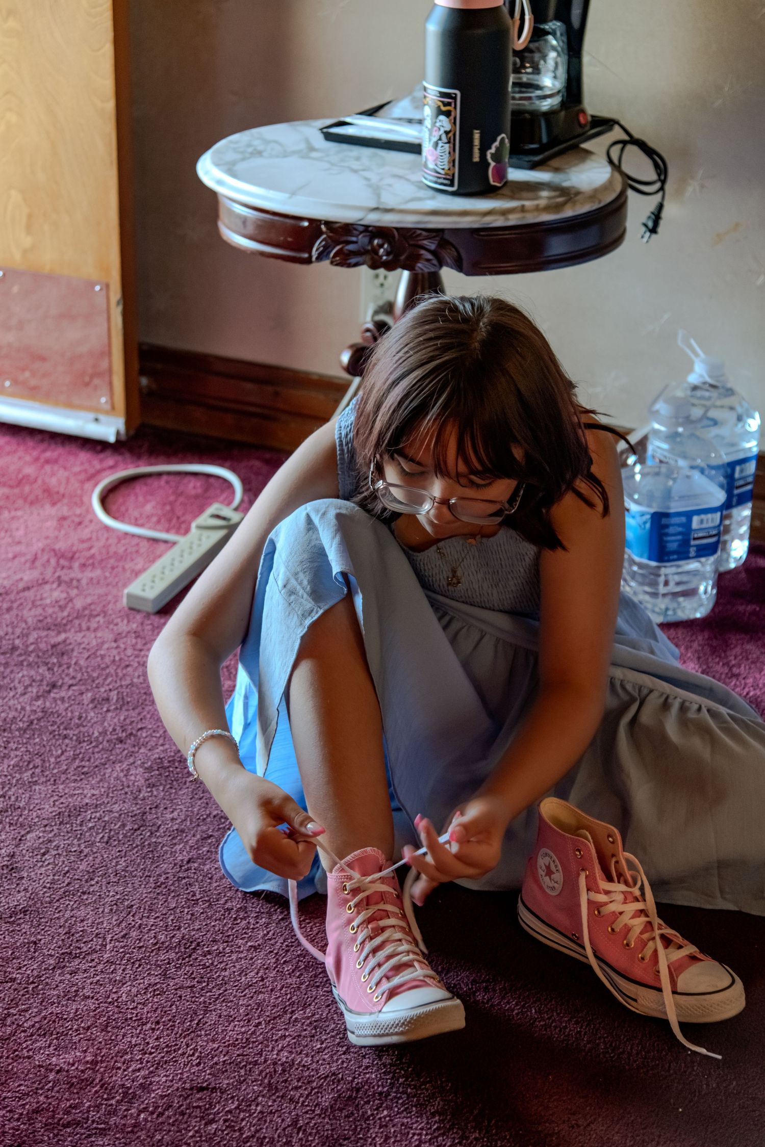 Young girl in blue dress and glasses tying pink Converse sneakers on red carpet floor.