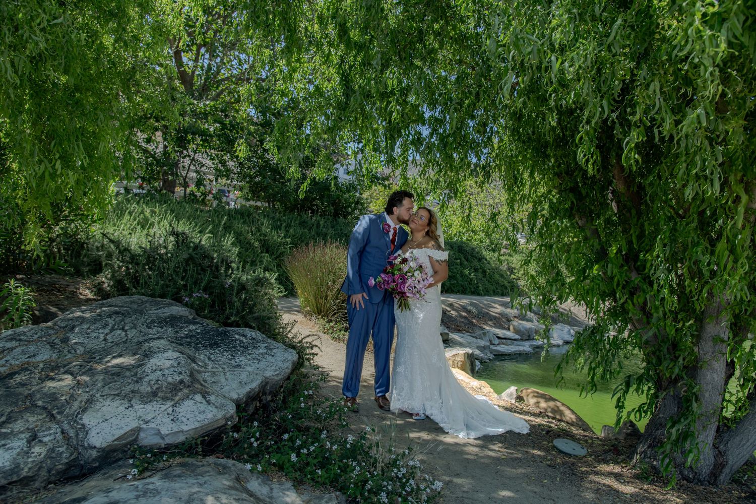 A couple stands on a large rock surrounded by lush willow trees in a romantic outdoor garden setting.