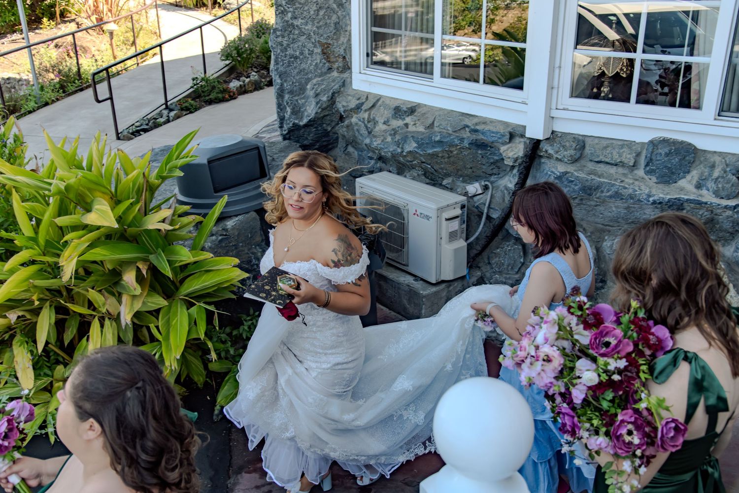 Bride in white gown sitting outdoors with bridesmaids holding colorful bouquets near stone building.