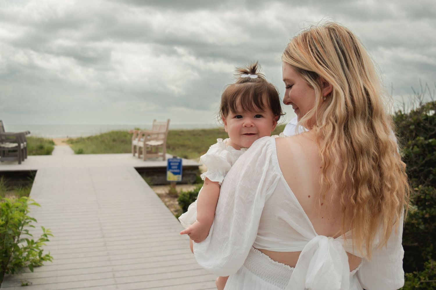 A mother in a white dress holds her baby girl on a beach boardwalk under cloudy skies.