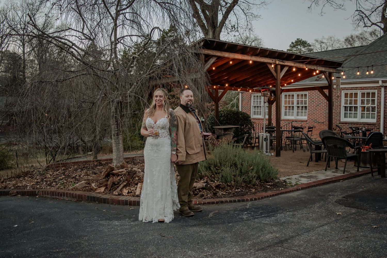 A wedding couple stands together at an outdoor venue with rustic barn and string lights at dusk during ceremony.