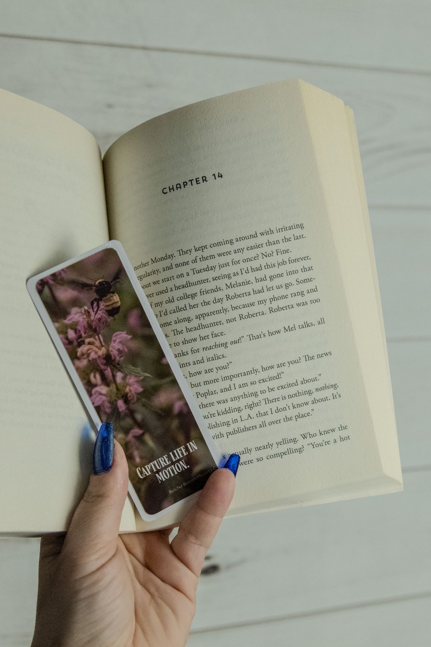 A hand with blue nail polish holds a phone displaying pink flowers against an open book showing Chapter 14.