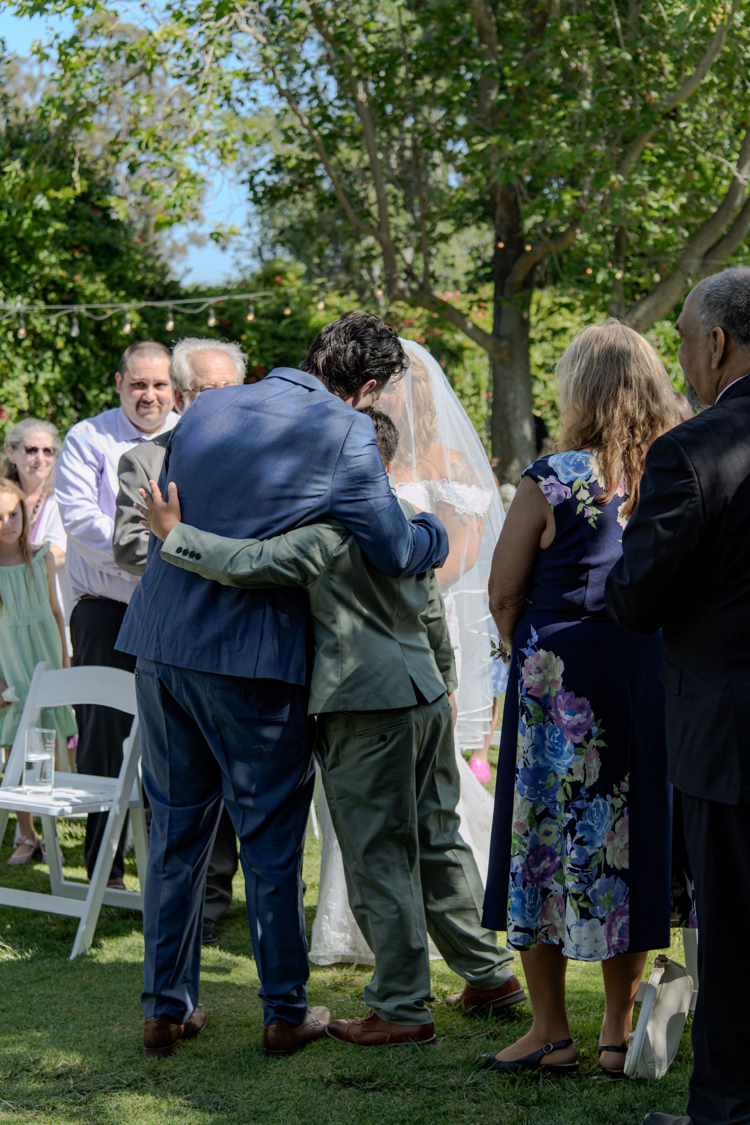 Groom embraces man in blue suit at outdoor wedding ceremony as bride in veil stands nearby.