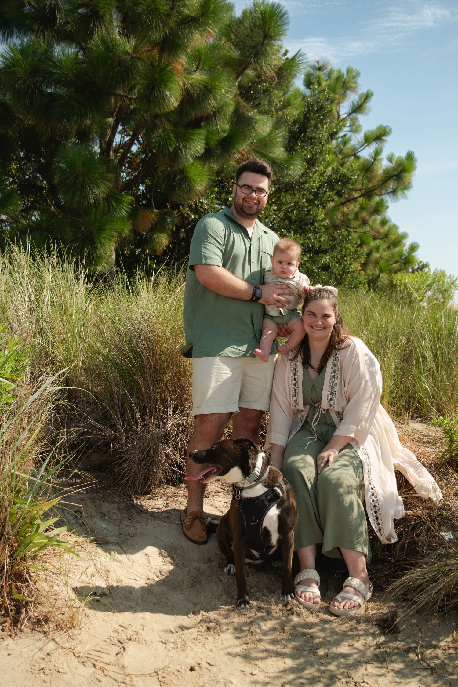 Family with baby and boxer dog posing on sandy path among dune grass and pine trees on sunny day.