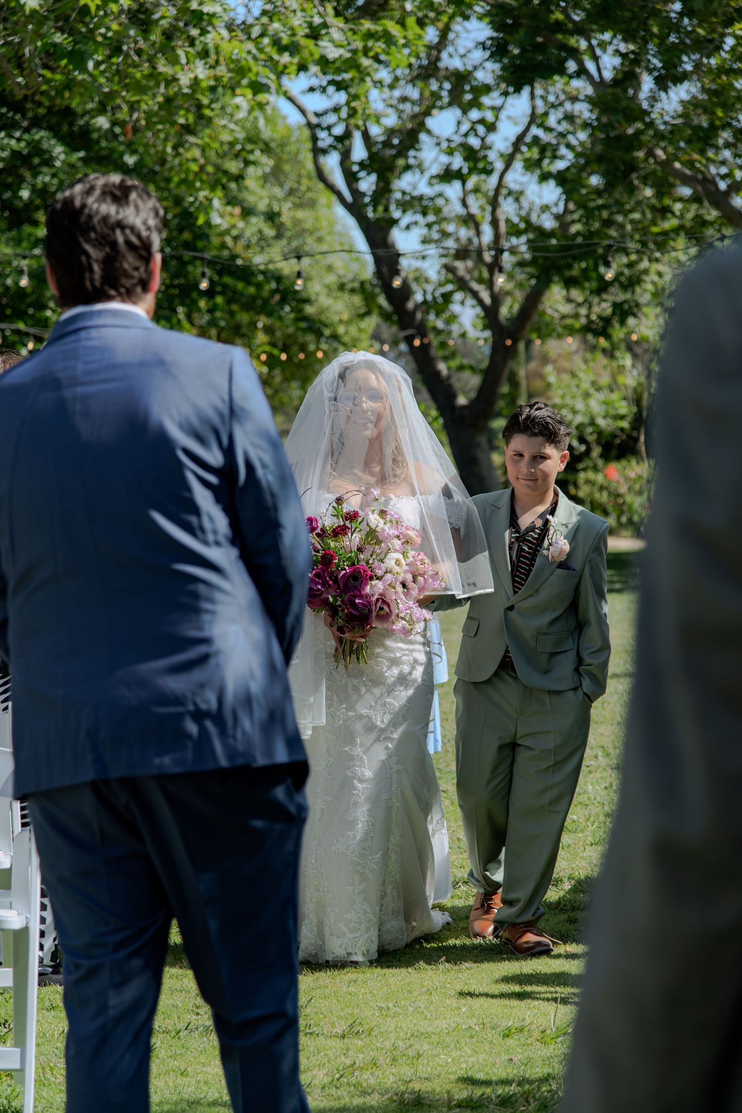 A wedding party stands in a garden setting with bridesmaids in lavender dresses and groomsmen in navy suits.