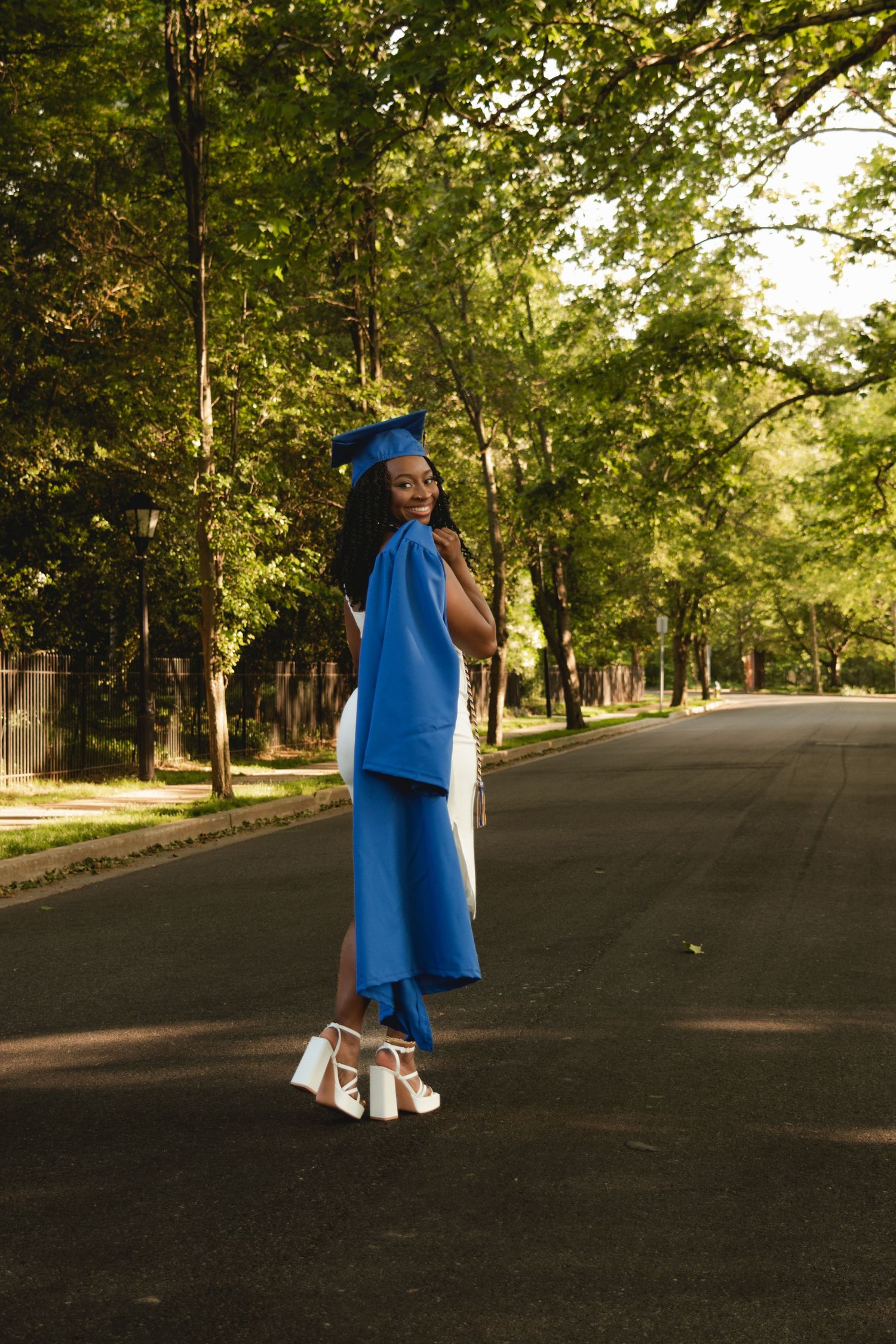 A person wearing blue graduation regalia and white dress walks through a tree-lined pathway as sunlight filters through foliage.