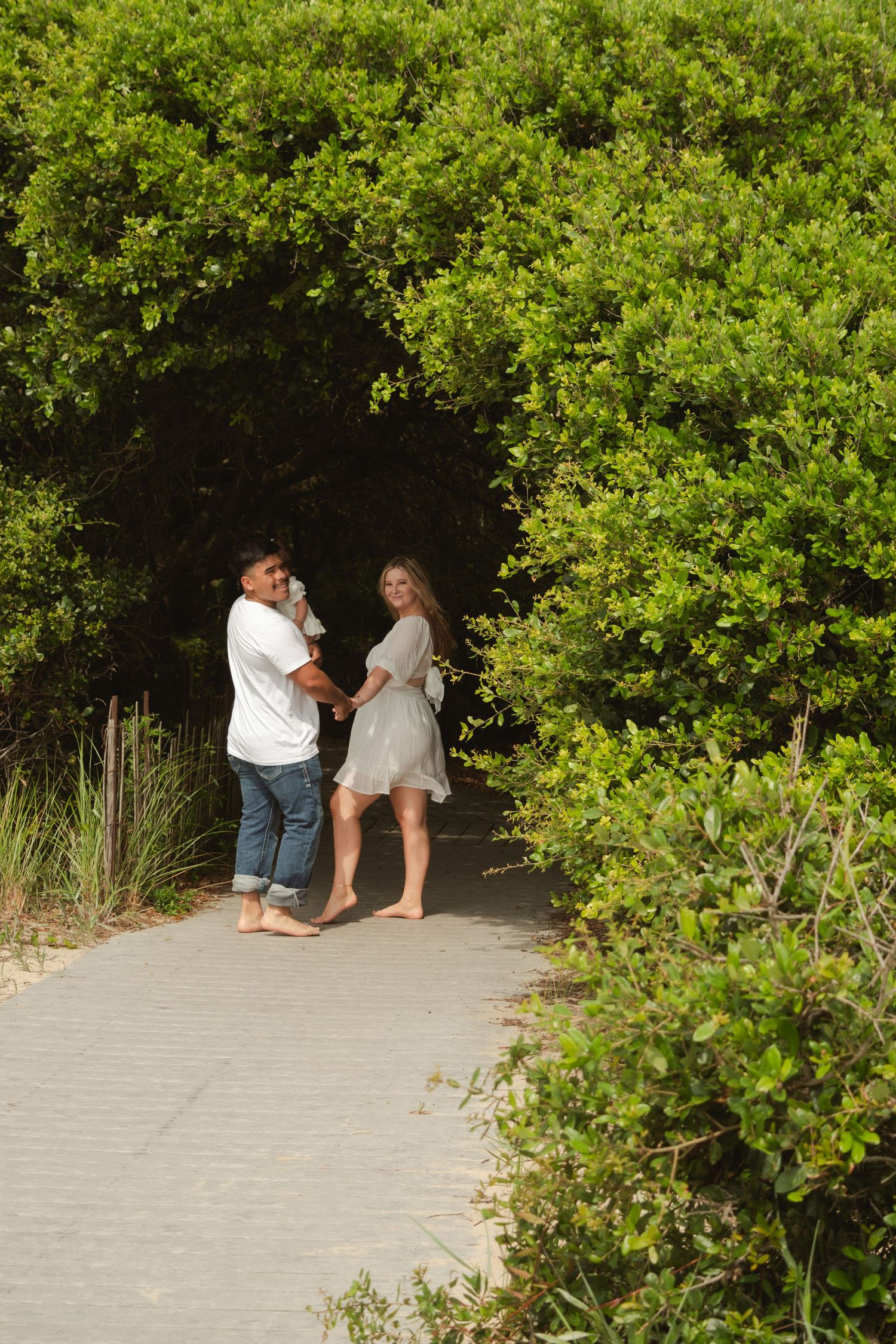 A couple walks hand-in-hand along a boardwalk path surrounded by lush green foliage, man carrying a small child.