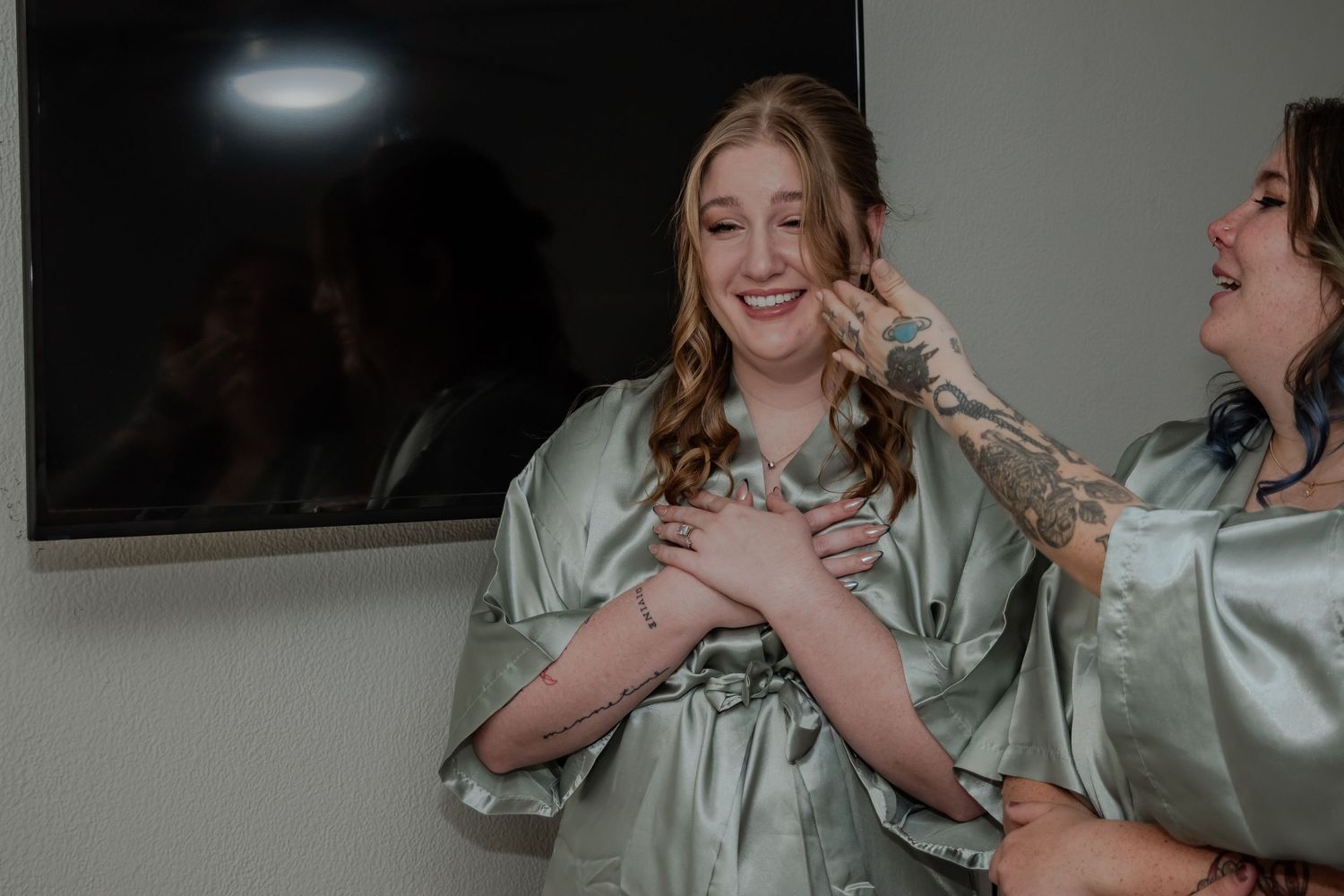 Group of friends share joyful laughter while wearing matching sage green satin robes during a celebration.