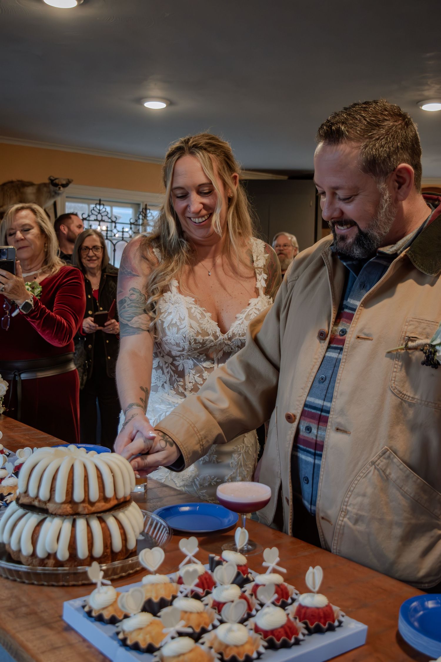 A couple cuts into a tiered Bundt cake while party guests gather around at a reception celebration.