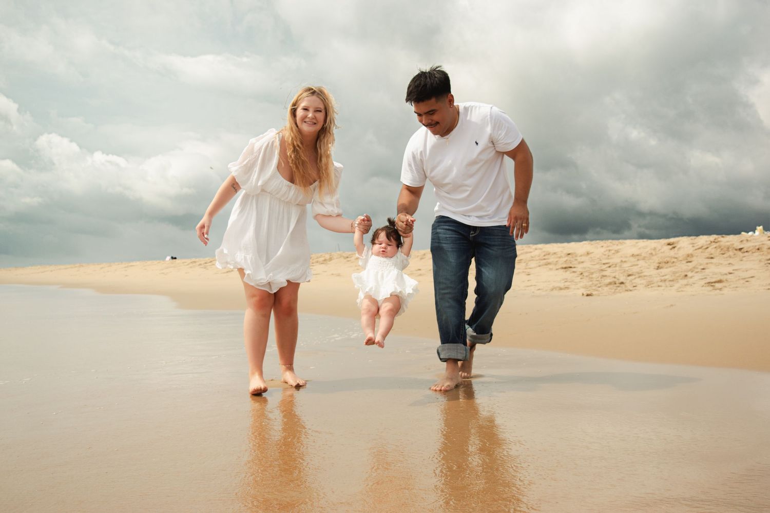 Family of three enjoying a beach walk, parents swinging toddler between them on a cloudy sandy shore.