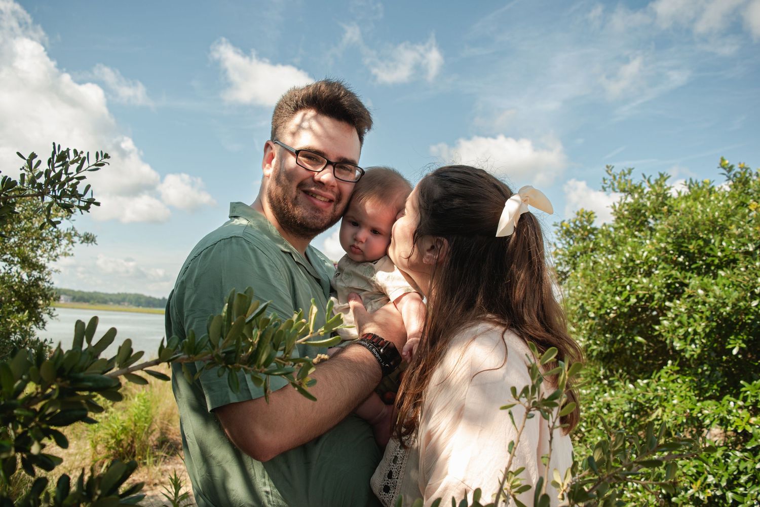 A young family with a baby poses outdoors near a lake surrounded by lush green shrubs under a blue sky.