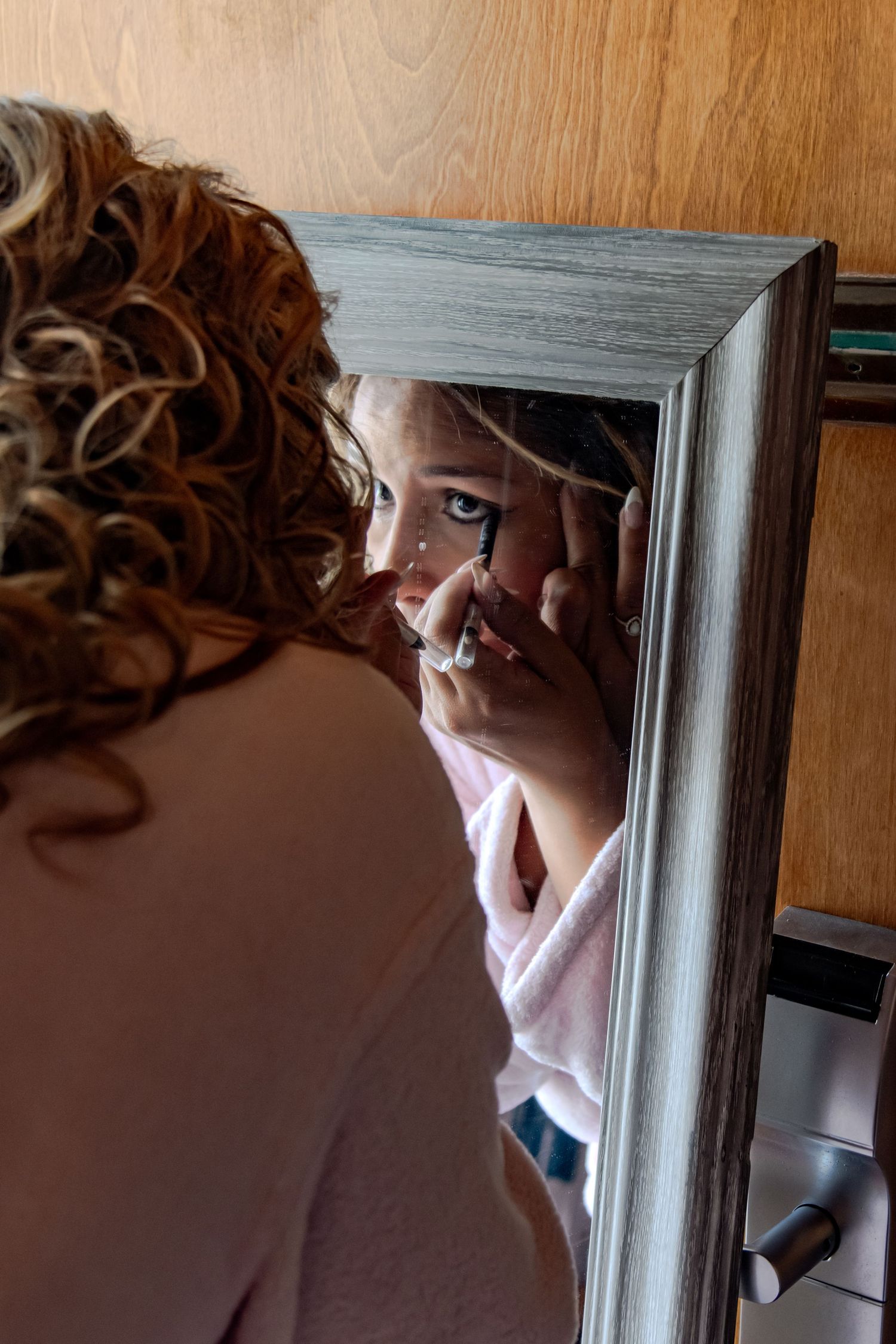Woman with curly hair applying eye makeup while looking into a gray wooden framed mirror.